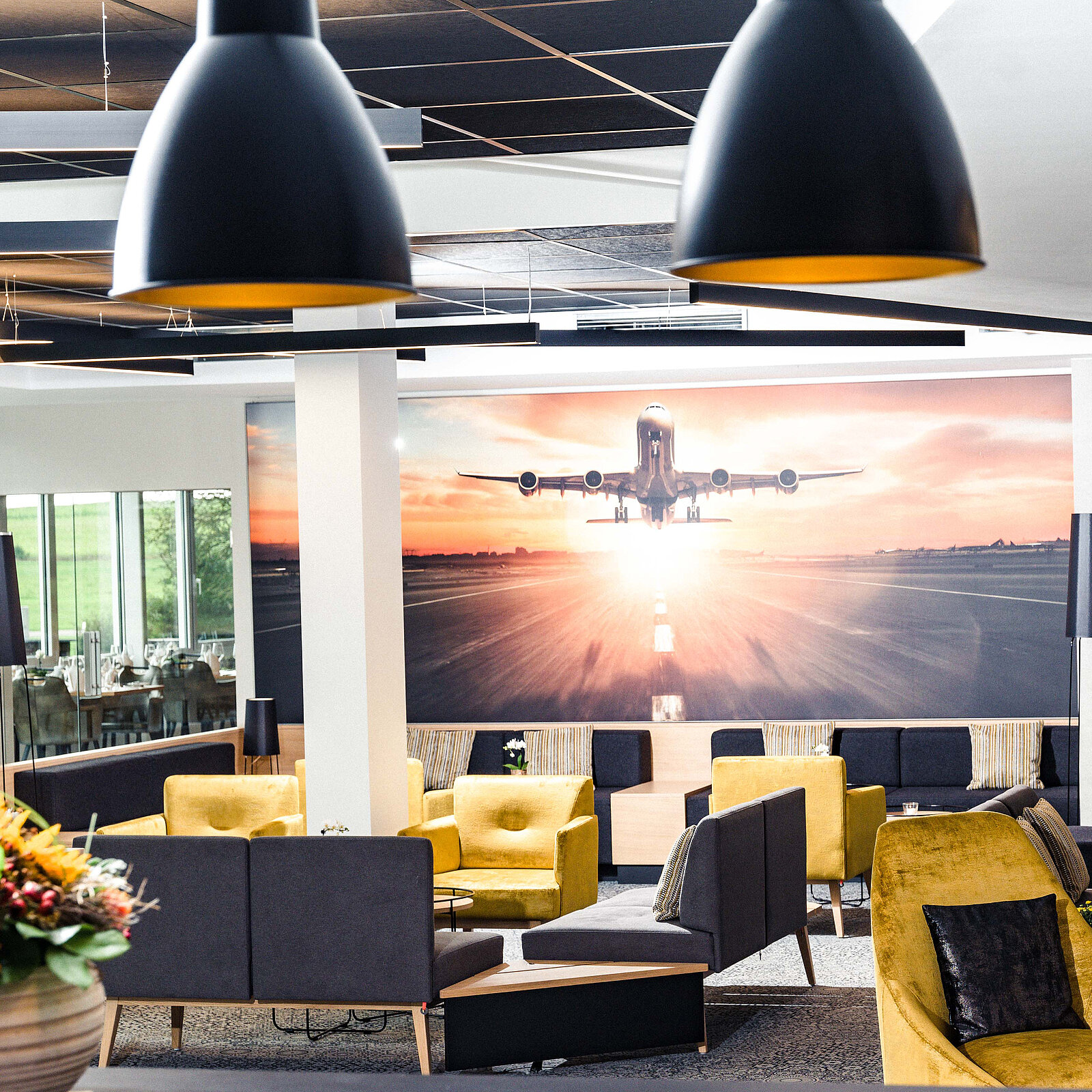 Modern lounge with yellow and gray seating, large airplane mural, pendant lights, and a window view of greenery.