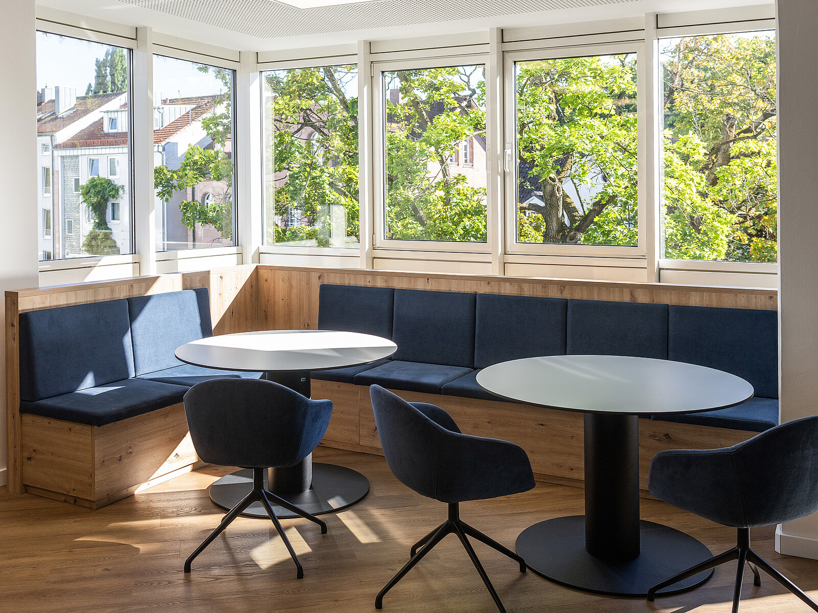 Moderne Sitzecke mit blauen Polstern, zwei runden Tischen und passenden Stühlen. Ein großer Fensterbereich bietet viel Tageslicht und Aussicht auf Bäume und Gebäude.