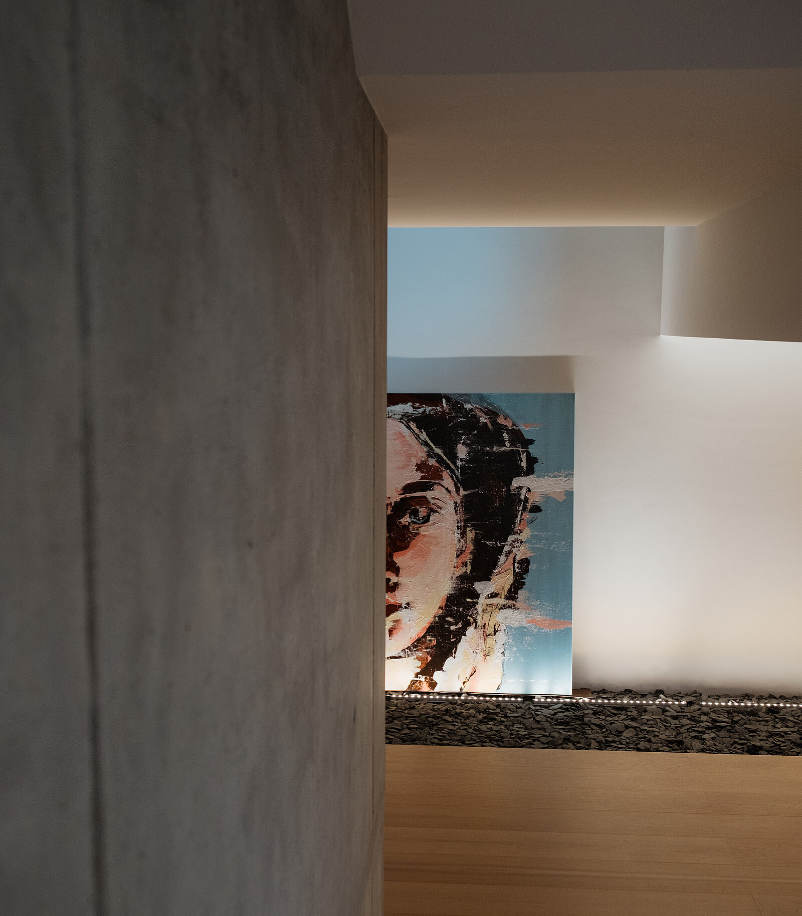 Nahaufnahme eines modernen Interieurs mit einer Teilwand und einem sichtbaren Kunstwerk. Das Kunstwerk zeigt das Gesicht einer Frau, das mit abstrakten, farbenfrohen Pinselstrichen vor einem blauen Hintergrund dargestellt ist, in einem gut beleuchteten, minimalistischen Raum.