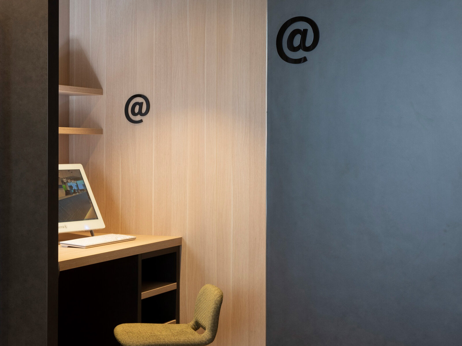 Minimalist workspace with a computer on a wooden desk, a green chair, and two black "@" symbols on light wood and dark gray walls.