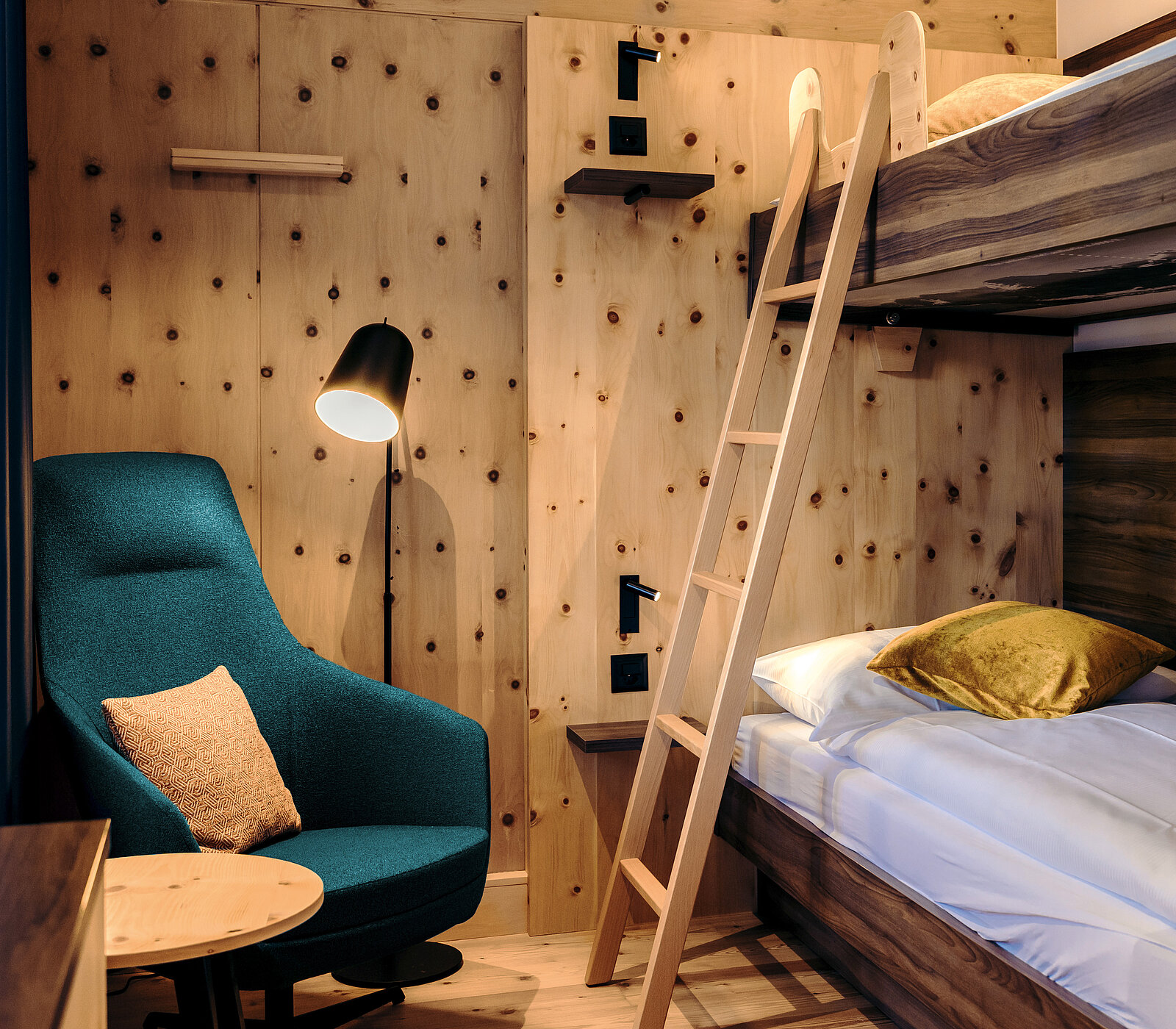 A cozy room featuring a blue armchair with a cushion, a small side table, and a ladder leading to a bunk bed. The walls are wooden with a natural finish, creating a warm atmosphere. The space is illuminated by a stylish floor lamp.