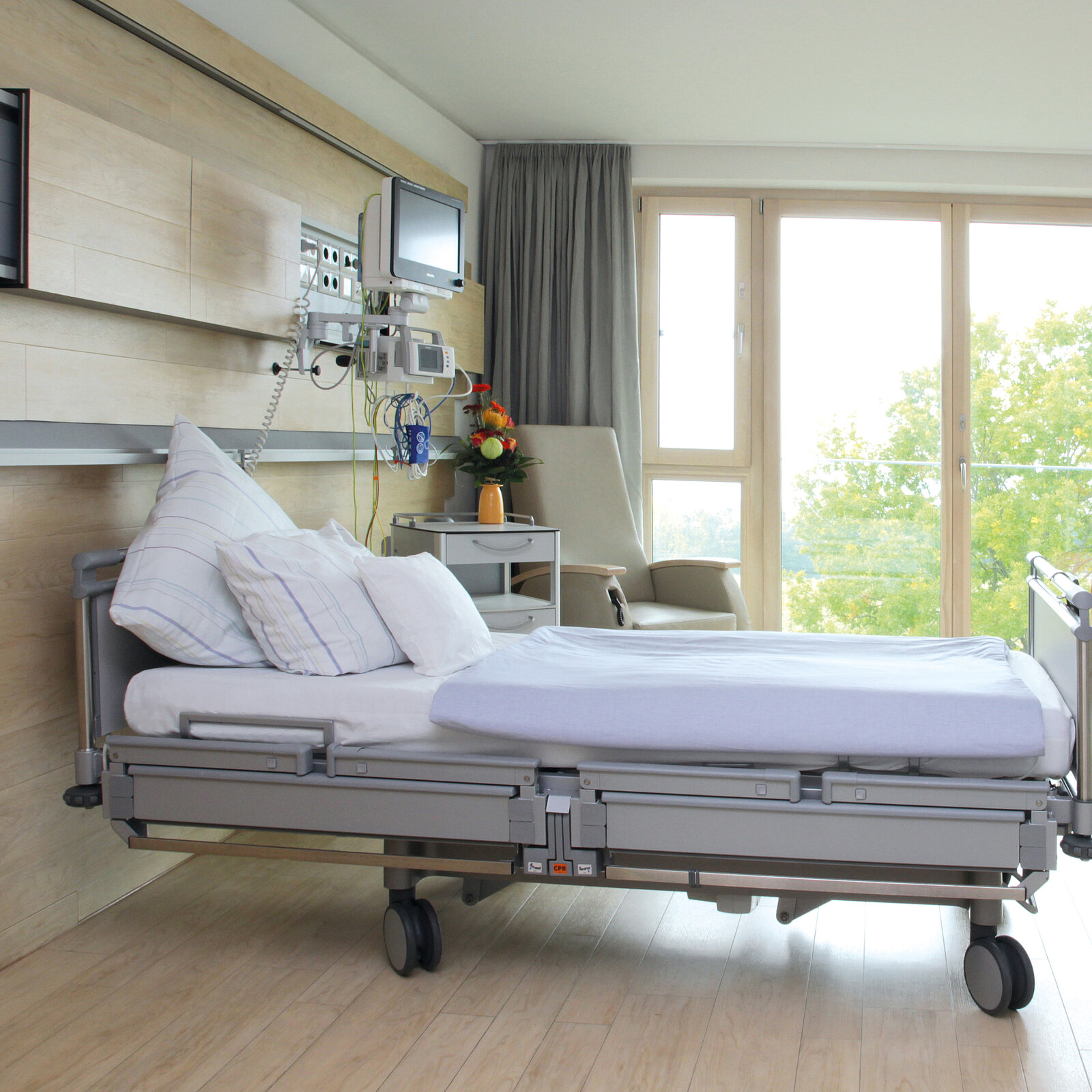Hospital room with an empty adjustable bed, medical equipment, a chair, and a window with a view of trees.