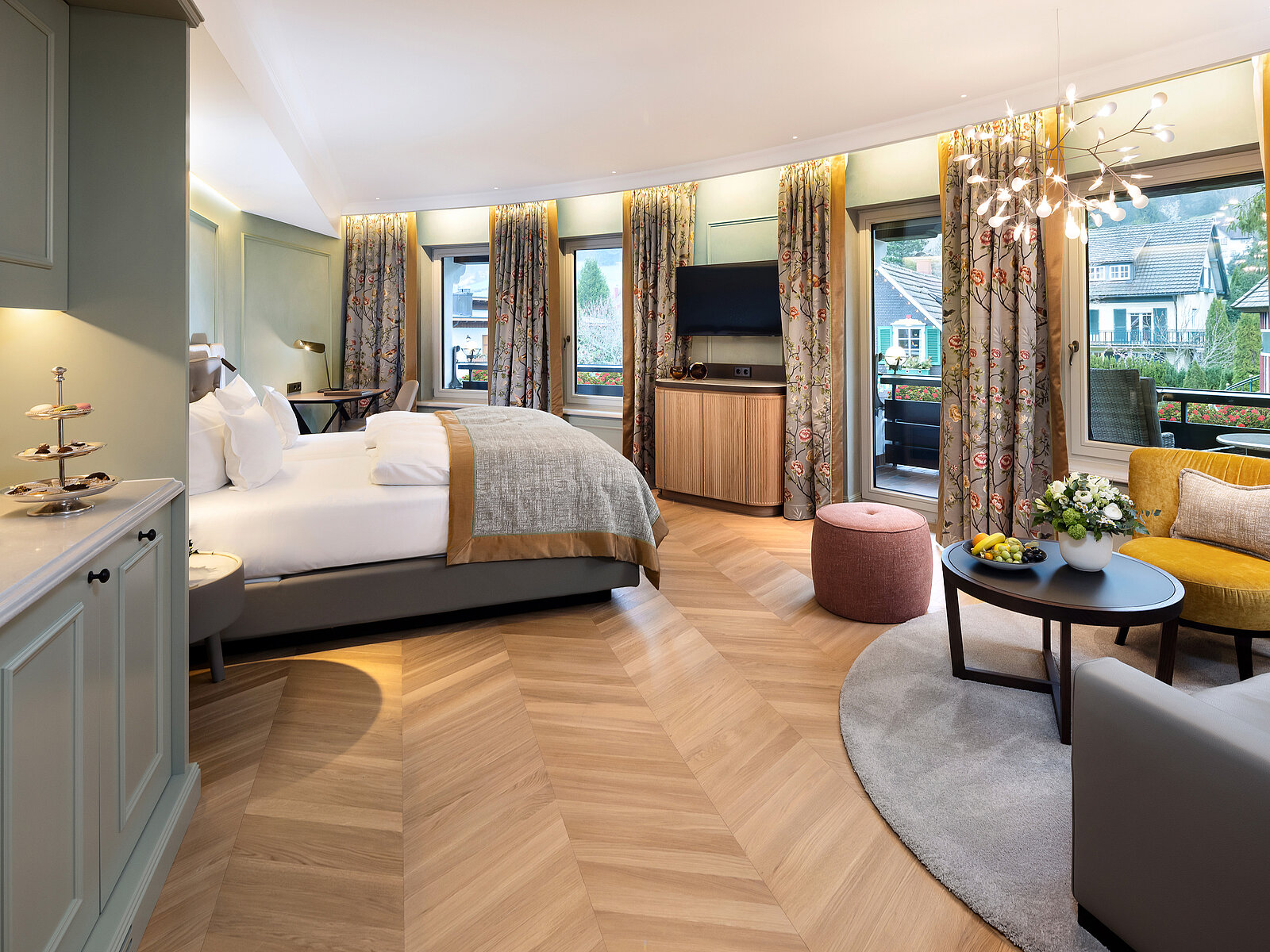 Helles und elegantes Schlafzimmer mit großem Bett, Flachbildfernseher, und Zugang zu einem Balkon durch große Fenster. Der Raum ist mit Holzboden, einem runden Teppich und stilvollen Möbeln ausgestattet. Dezente Beleuchtung und Vorhänge schaffen eine gemütliche Atmosphäre.