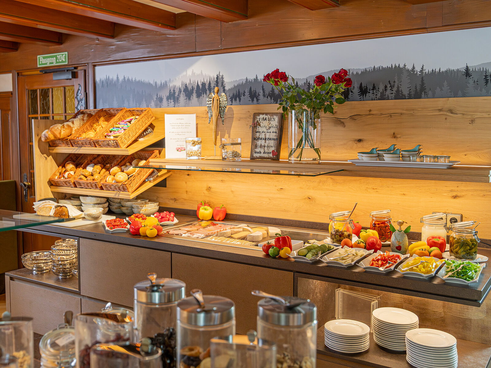Das Bild zeigt ein vielfältiges Frühstücksbuffet mit Brotkörben, frischen Früchten, Aufschnitt, Gemüse und verschiedenen Gläsern mit Marmeladen. Dekoriert mit roten Rosen und einer Berglandschaft im Hintergrund bietet es eine einladende Atmosphäre.