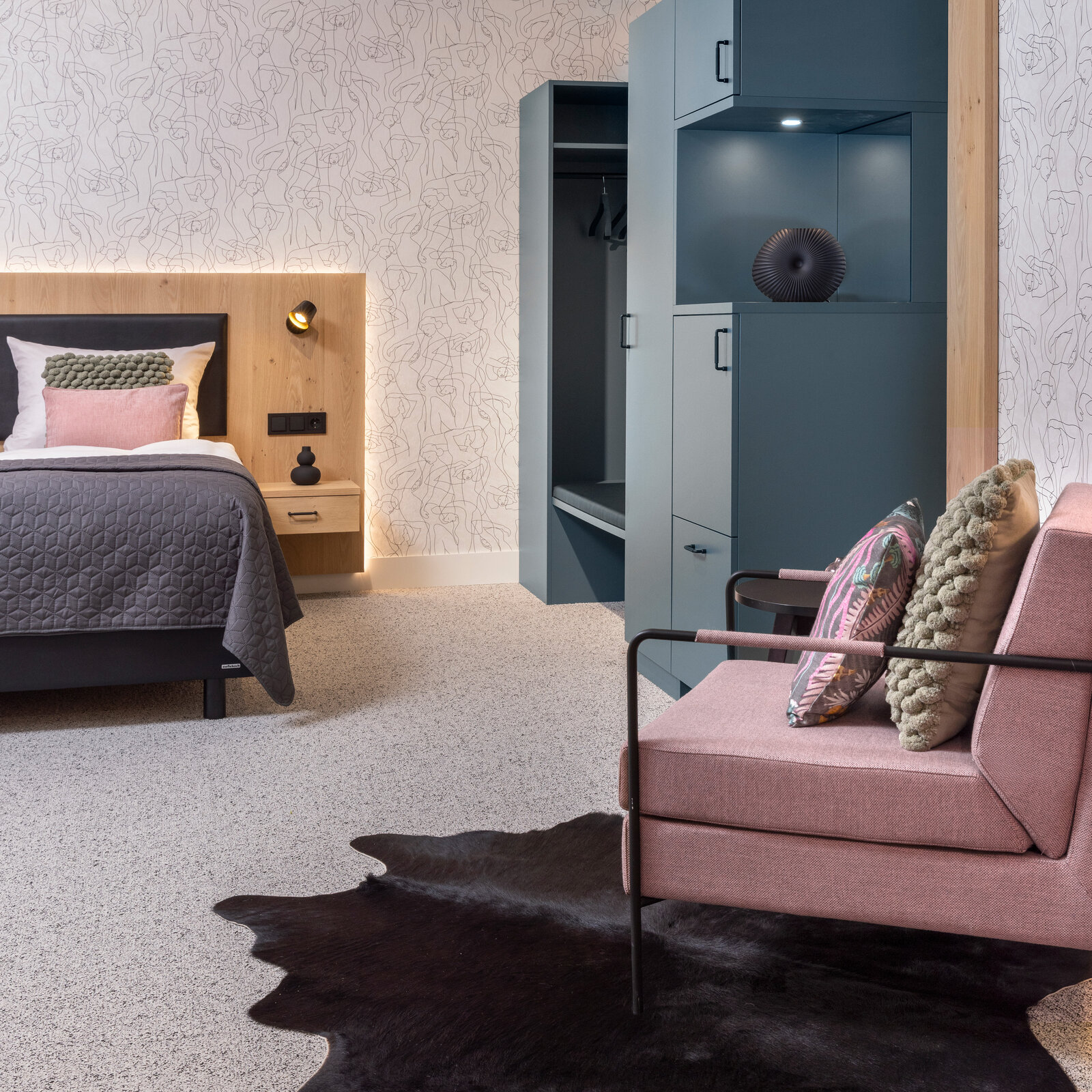 zieflekoch musterzimmer zenit, modernes Schlafzimmer mit einem Boxspring-Doppelbett mit Rückwand in Holzoptik, einem rosafarbenen Sessel, einem Schrank in gedecktem Grün und einer abstrakten Motivtapete, der Teppichboden und ein dunkler Teppich verleihen dem Raum Wärme.