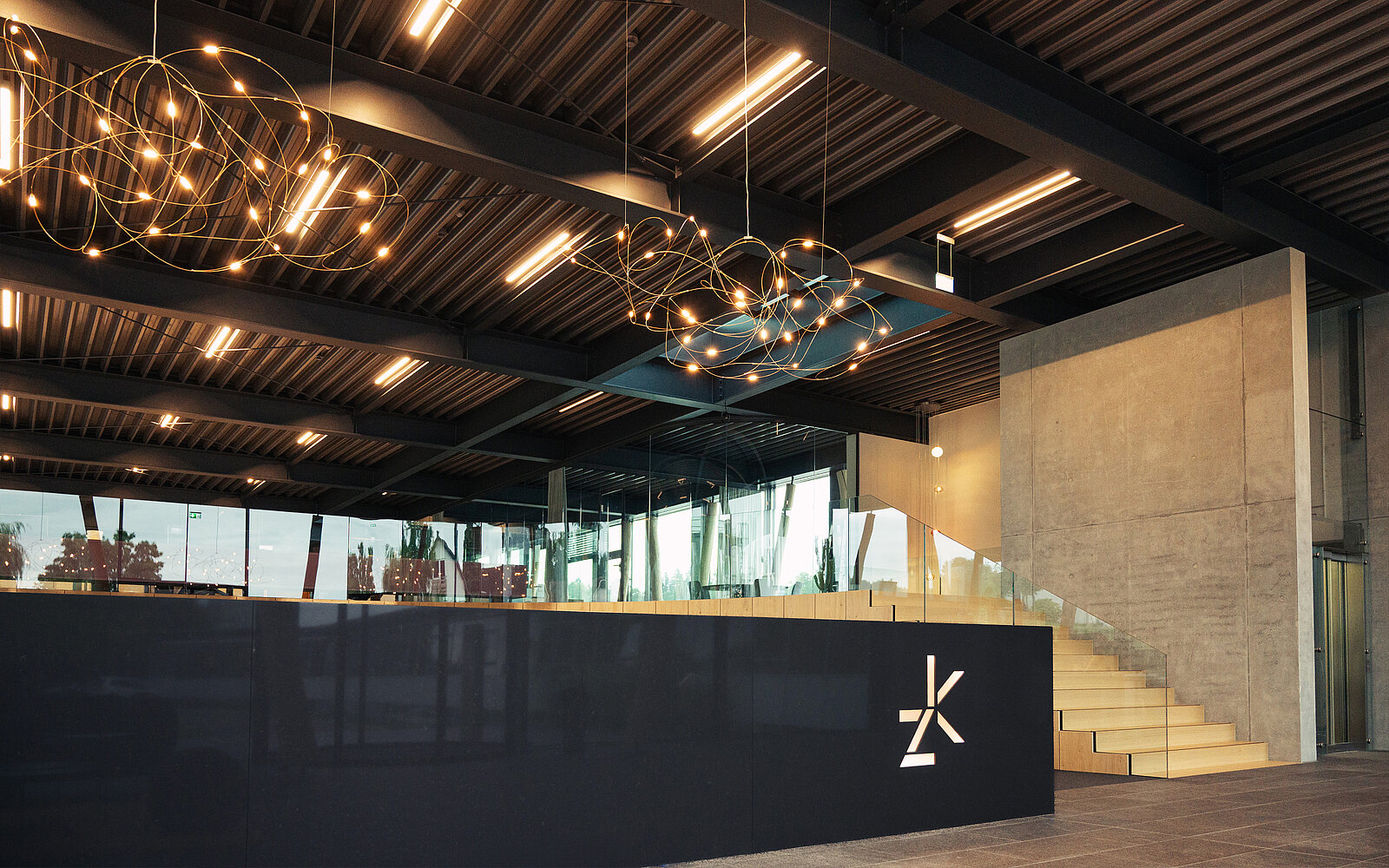 Die elegante Lobby verfügt über eine moderne Rezeption mit minimalistischem Design, die mit einem eleganten Logo verziert ist. Über ihr hängen moderne Leuchten von der Decke und beleuchten den Raum, während eine Treppe zu einer oberen Etage führt, die durch große Glaswände im Hintergrund sichtbar ist.