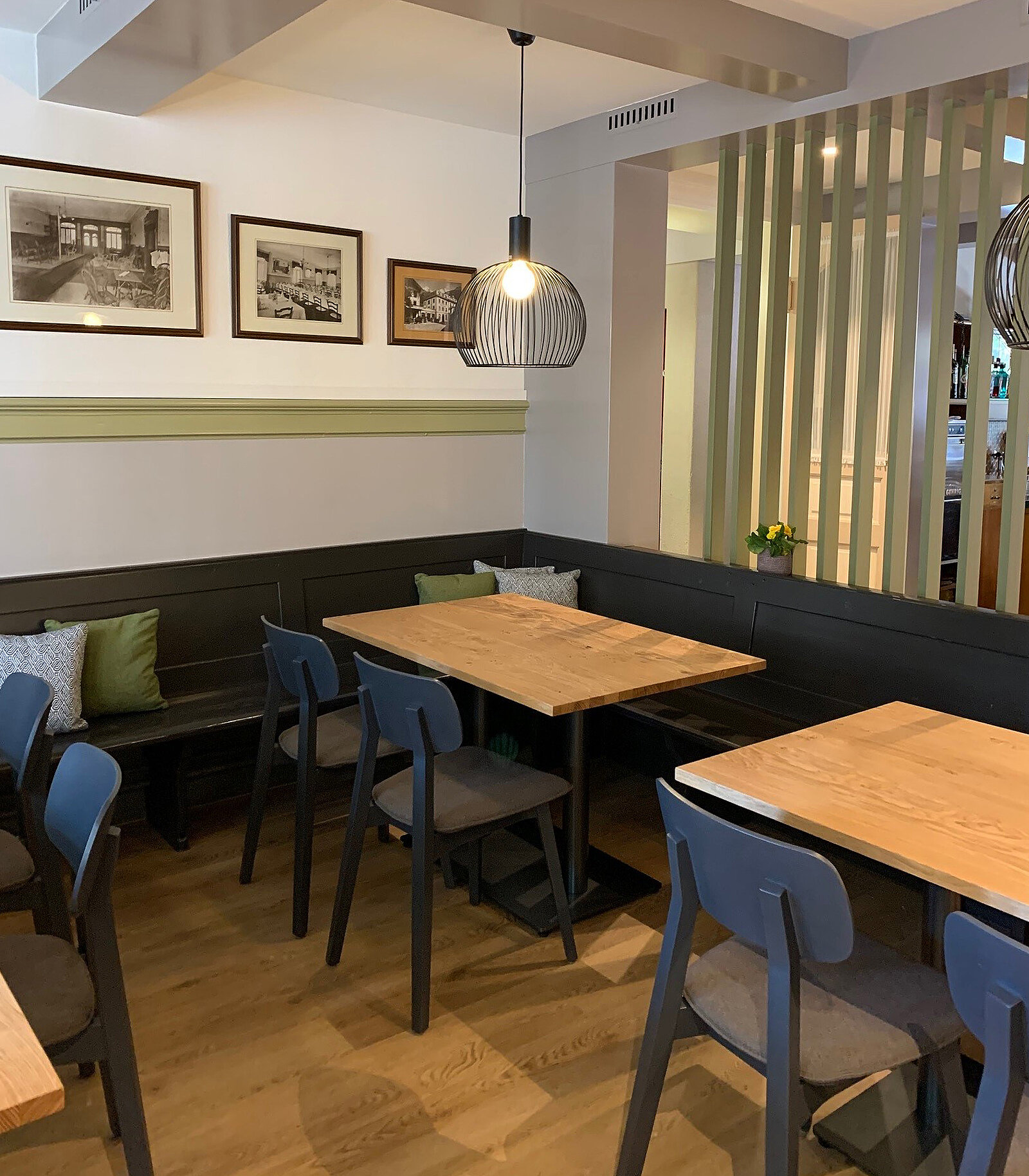 Ein Restaurantbereich mit Holzböden und modernen Tischen. An der Wand hängen gerahmte Bilder. Die Sitzbänke sind mit Kissen ausgestattet. Eine Pendelleuchte hängt über einem der Tische, während im Hintergrund ein heller, abgeteilter Bereich sichtbar ist.