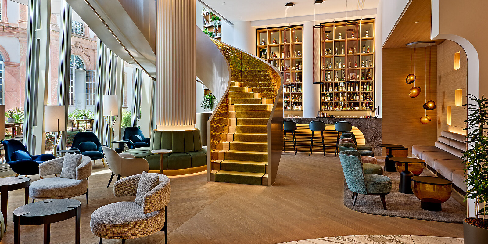 Eine stilvolle Hotel-Lobby mit geschwungener Treppe, modernen Sesseln und einem beleuchteten Regal mit Flaschen. Elegante Beleuchtung und große Fenster schaffen eine einladende Atmosphäre.