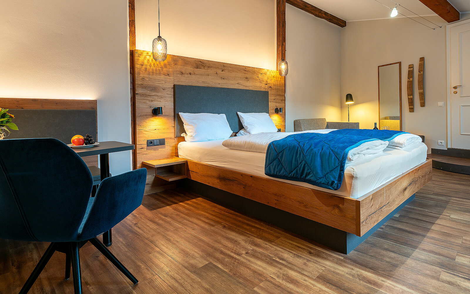 Ein modernes Hotelzimmer mit einem großen Doppelbett, weißer Bettwäsche und einer blauen Decke. Angeordnet sind ein bequemer Sessel und ein kleiner Tisch. Die Wände sind hell, das Zimmer hat Holzboden und warmes Licht von Pendelleuchten.
