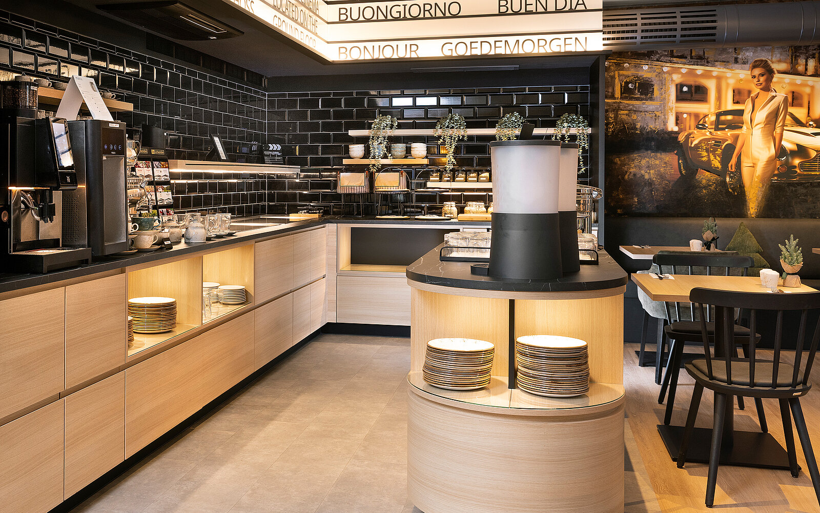 A modern café interior features dark tiled walls and wooden accents. The counter showcases various beverages and snacks in stylish displays. Soft lighting highlights a round table with chairs, contributing to a cozy and inviting atmosphere. A framed artwork adds a decorative touch to the space.