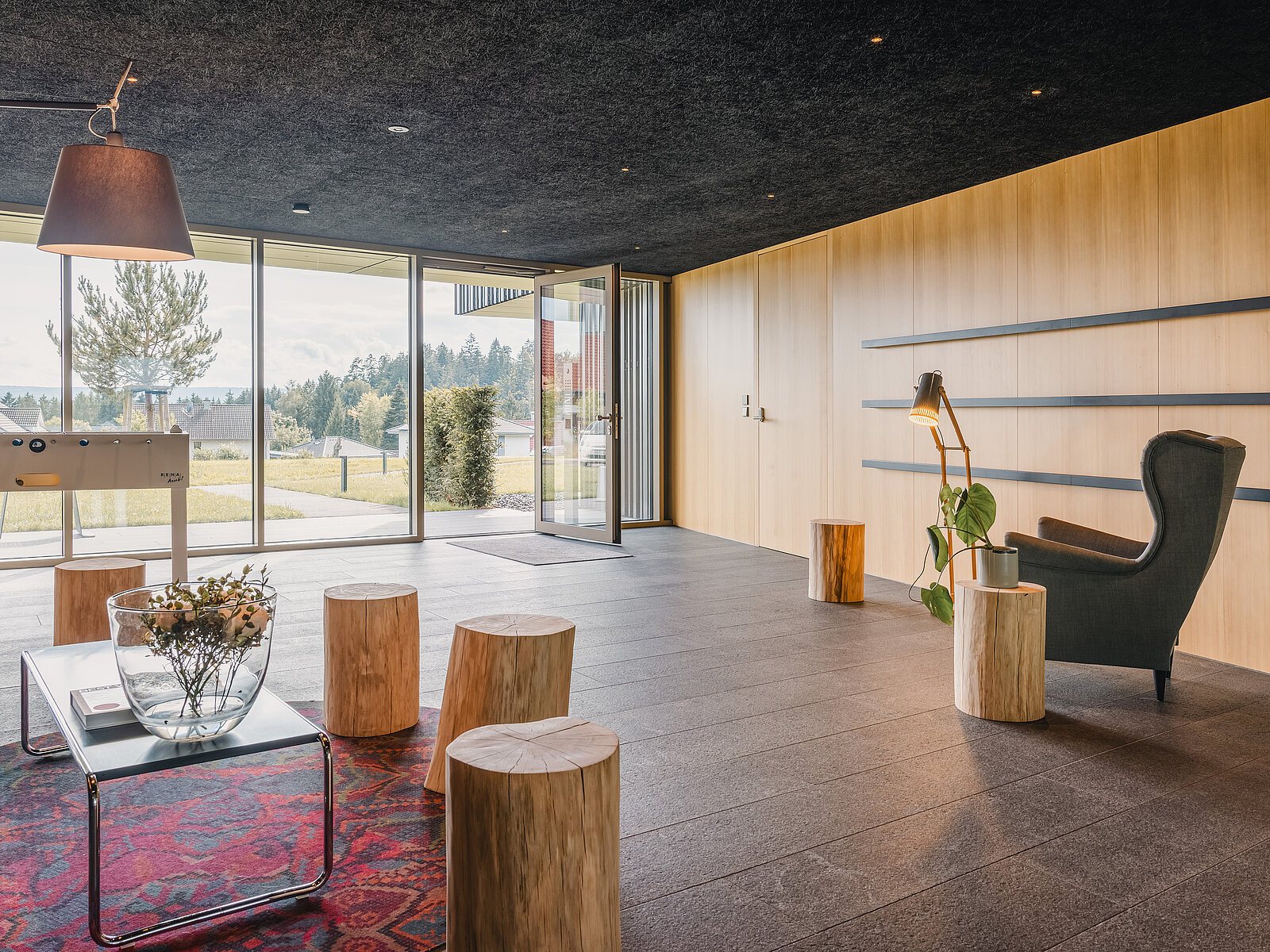 Moderner Raum mit Holzelementen: Holzstümpfe als Sitzhocker, ein schwarzer Sessel und eine Stehlampe. Große Glasfront gibt Blick auf grüne Landschaft frei.