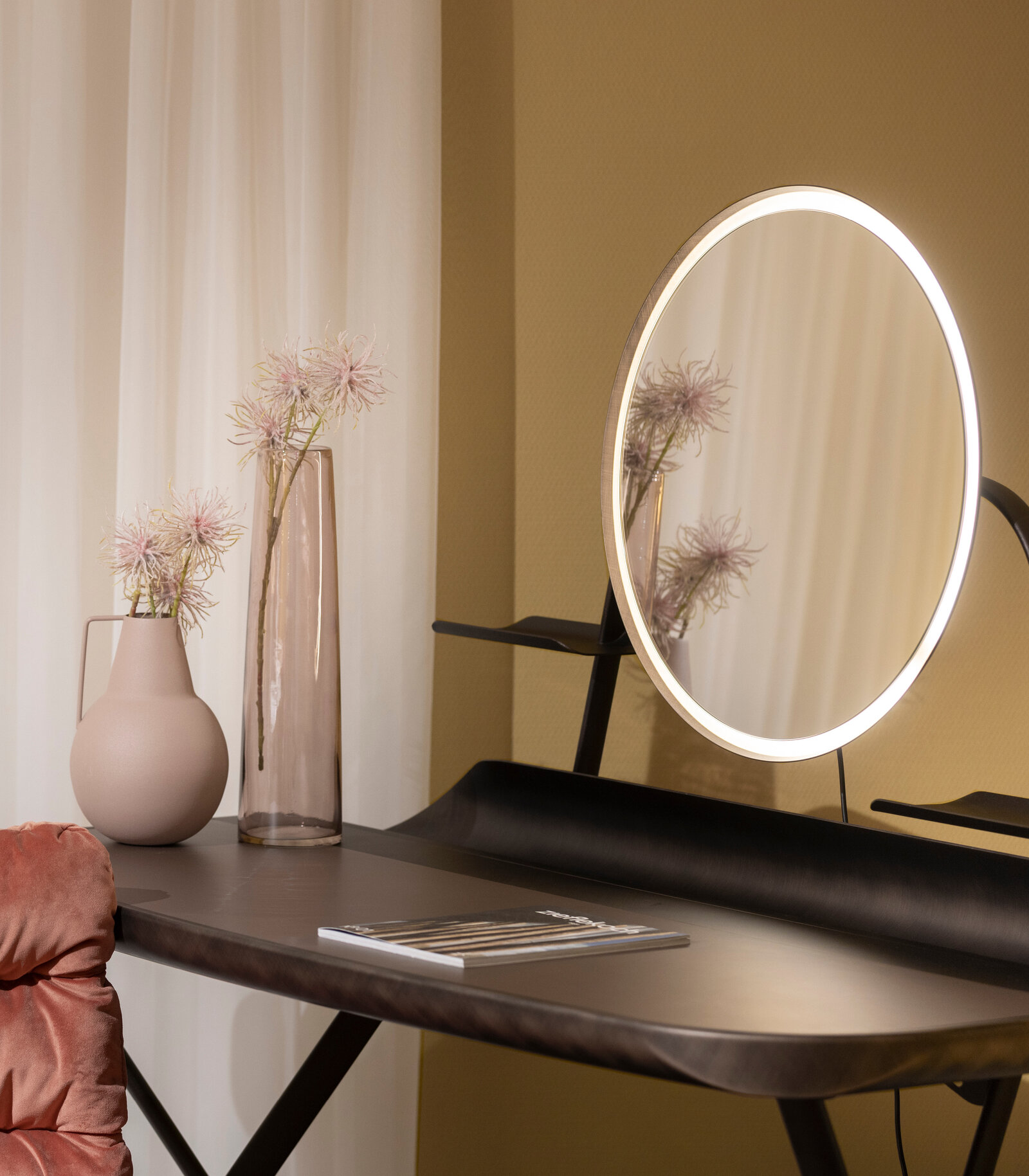 A vanity table with a round, illuminated mirror, a pink vase, a tall glass vase with flowers, and a magazine, set against beige walls and white curtains.
