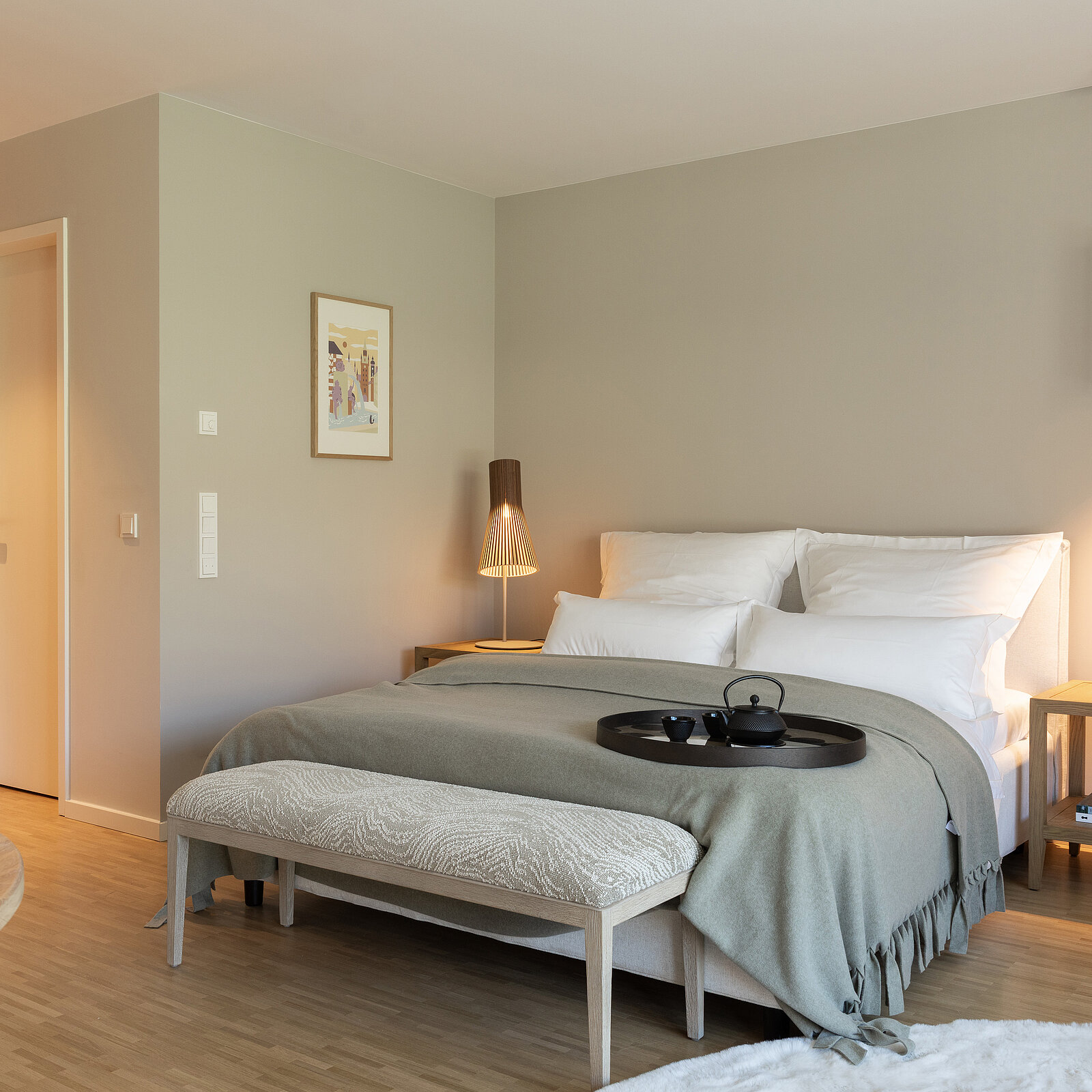 Ein stilvoll eingerichtetes Schlafzimmer mit einem großen Bett, weißer Bettwäsche, grauer Decke und einem Tablett mit Teekanne darauf. Neben dem Bett stehen Nachttische mit Lampen. Eine dezent beige Wand mit einem Bild vervollständigt das gemütliche Ambiente des Raumes.