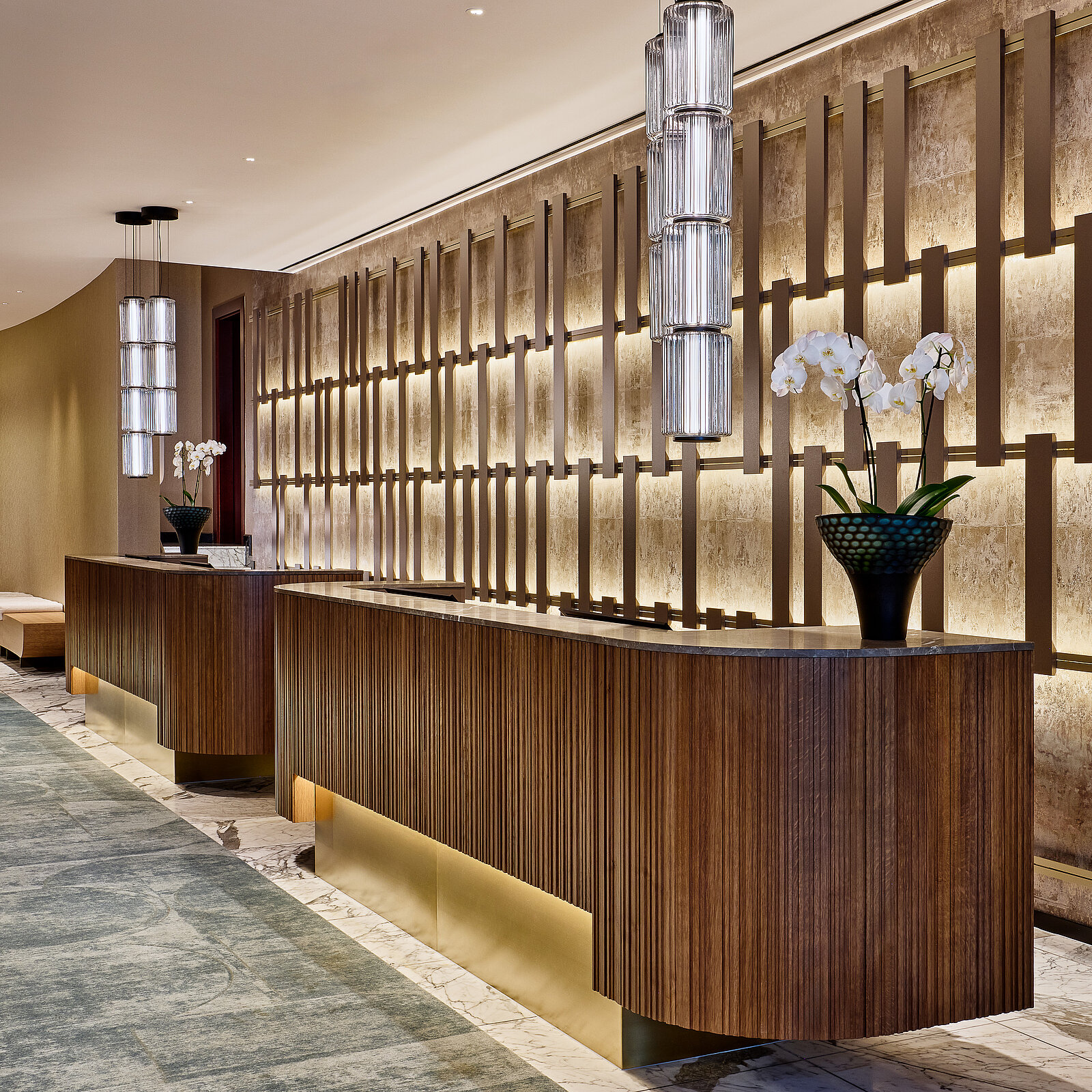 Der luxuriöse Empfangsbereich eines Hotels mit hölzernen Schreibtischen, modernen Lampen und einer dekorativen Wand. Orchideen in Vasen stehen auf den Tischen. Ein eleganter Teppich verläuft entlang des Bodens, der Raum wirkt stilvoll und einladend.