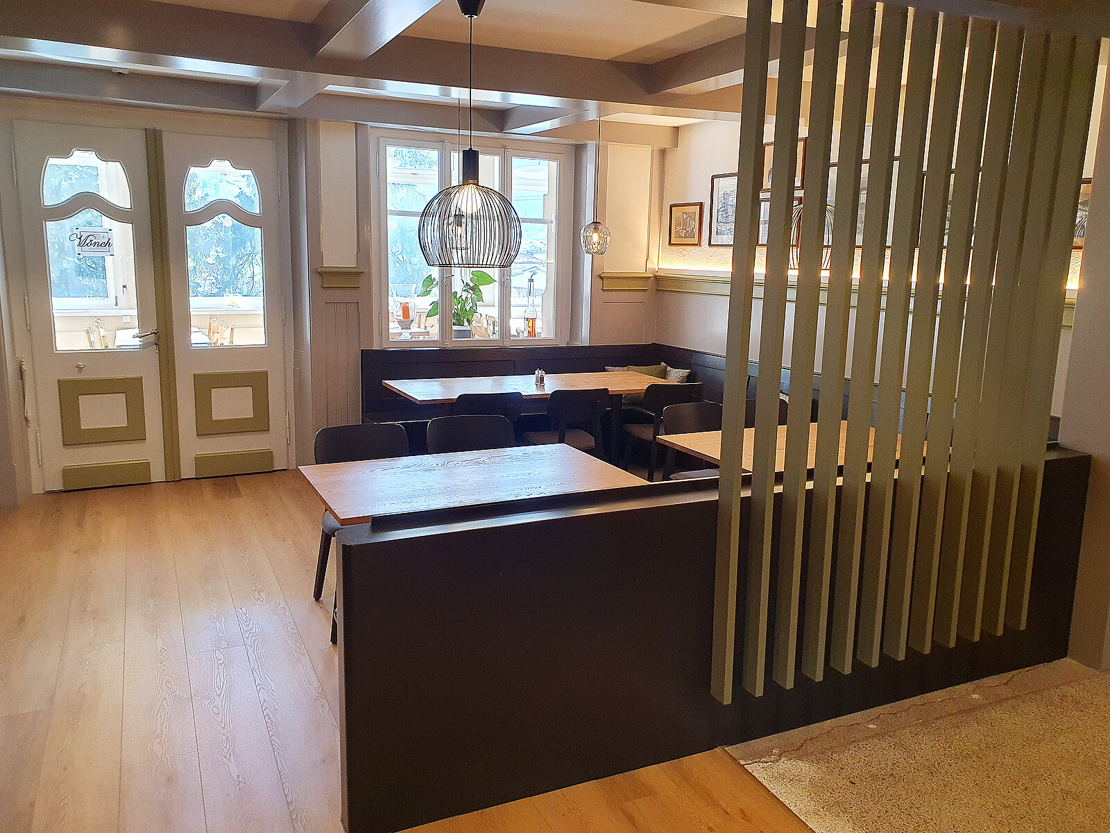 Ein gemütliches Restaurantinnere mit Holzboden und Trennwänden aus senkrechten Latten. Mehrere Tische und Stühle sind sichtbar. An der Wand hängen Bilder, und eine Pendelleuchte sorgt für angenehmes Licht. Im Hintergrund sind Fenstertüren, die auf eine andere Atmosphäre hinweisen.