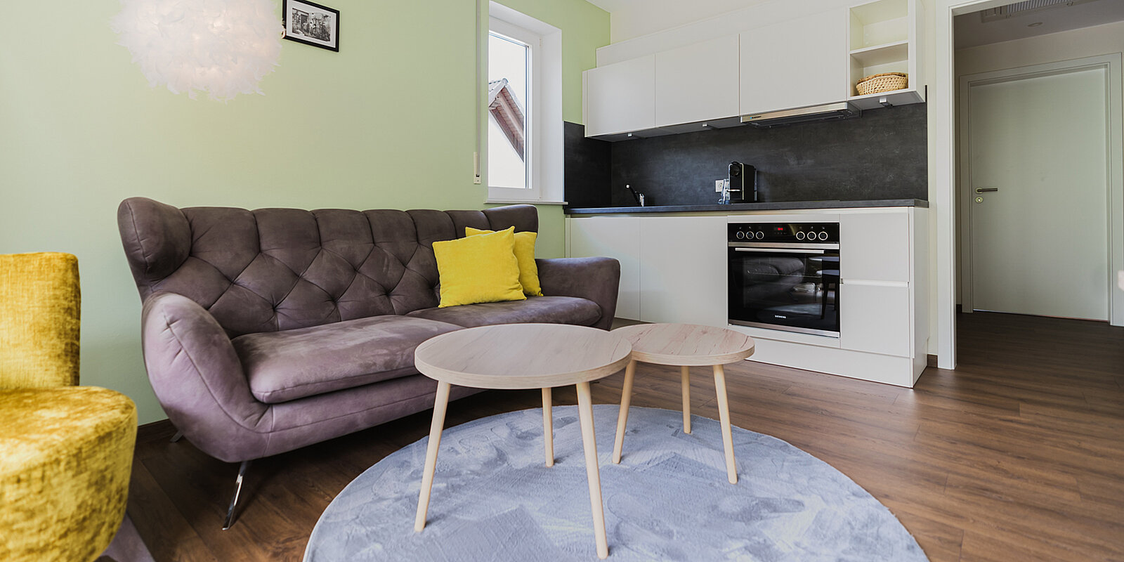 Gemütliches Wohnzimmer mit grauem Sofa, gelbem Kissen und zwei runden Holztischen auf einem runden, blauen Teppich. Im Hintergrund befindet sich eine moderne Küchenzeile mit Herd und dunkler Rückwand. Die Wände sind hellgrün, der Boden ist aus Holz.