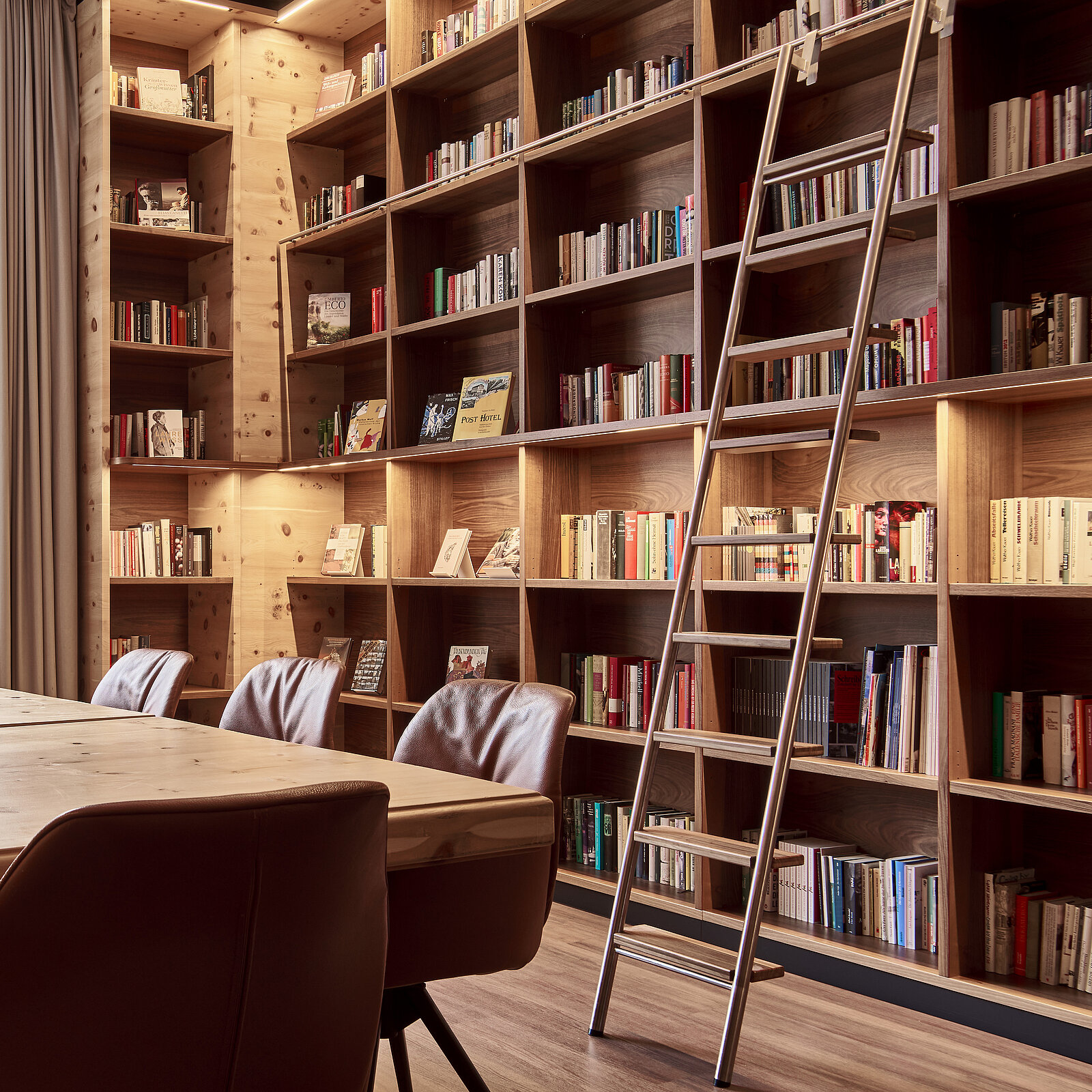 A cozy room featuring tall wooden bookshelves filled with books. A long table with leather chairs is in the foreground, with a ladder leaning against the shelves for accessing higher books. Soft lighting highlights the wood tones, creating a warm atmosphere.