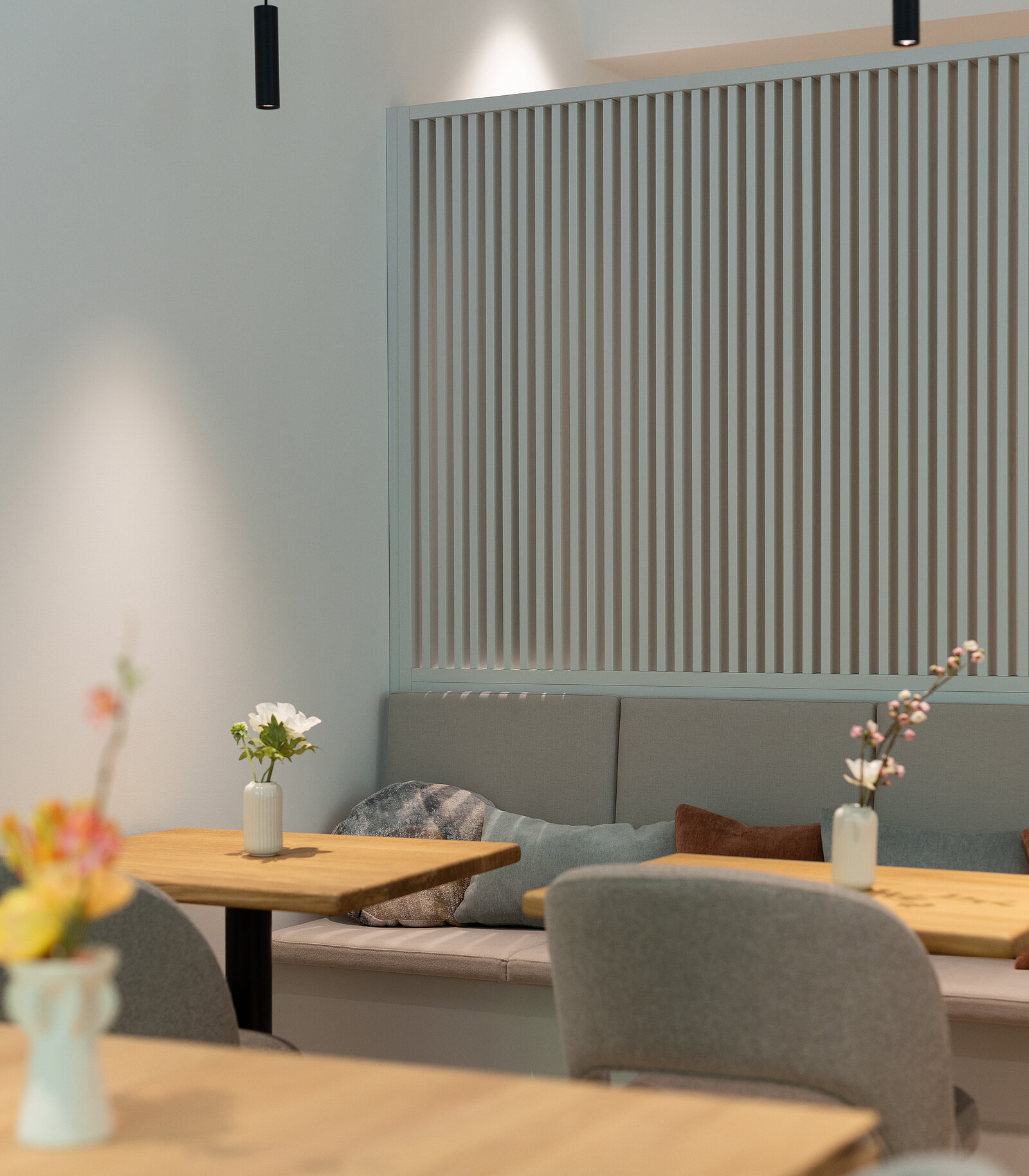 Ein Café mit modernen, hellen Holztischen und grauen Stühlen. Auf den Tischen stehen kleine Blumenvasen mit bunten Blumen. Im Hintergrund sieht man eine Sitzbank mit Kissen und eine dekorative Trennwand aus vertikalen Lamellen.