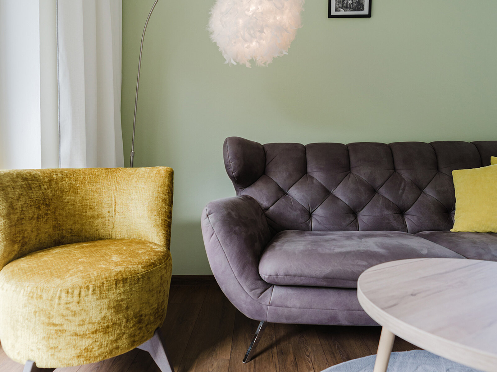 Gemütliches Wohnzimmer mit einem grauen, gesteppten Sofa und einem runden, goldgelben Sessel. Eine flauschige Hängelampe und ein kleiner runder Tisch ergänzen die Einrichtung. Die Wände sind hellgrün gestrichen.