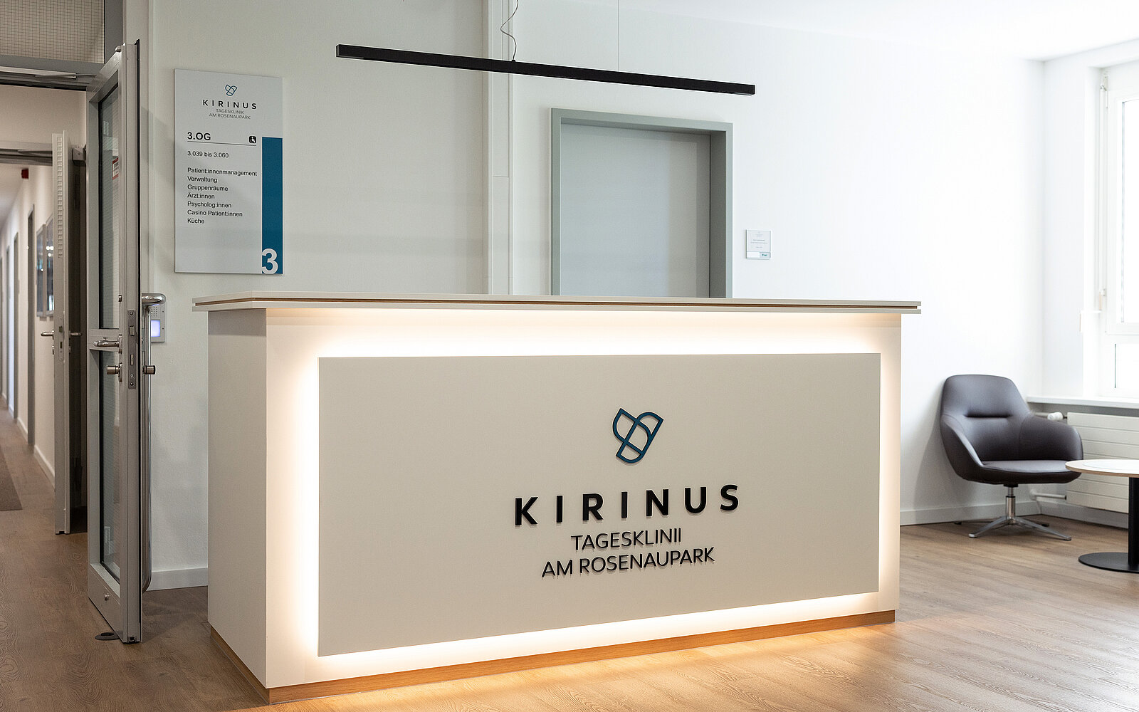 Empfangsbereich einer Klinik mit modernem Tresen. Auf dem Tresen steht "KIRINUS Tagesklinik am Rosenaupark". Der Raum wirkt hell und einladend, mit einem schwarzen Stuhl und minimalistischer Dekoration.