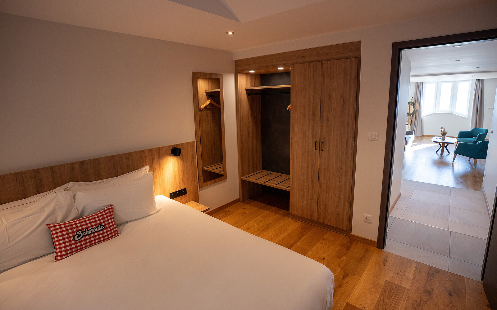 Modernes Hotelzimmer mit einem großen Bett, einem Holzkleiderschrank und einem angrenzenden Wohnbereich mit hellen Fenstern. Ein Kissen mit kariertem Muster ziert das Bett.