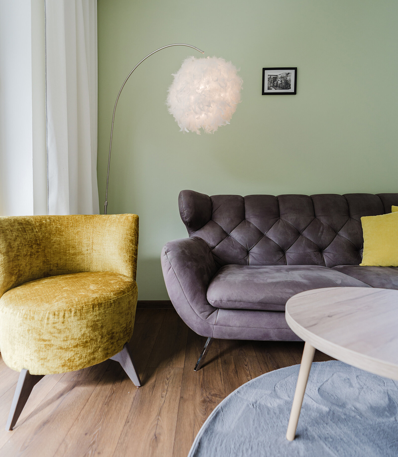 Ein Wohnzimmer mit einer grün gestrichenen Wand. Es enthält ein graues Sofa mit gelben Kissen, einen gelben Sessel, eine runde weiße Stehlampe und einen runden Holztisch auf einem grauen Teppich. Ein kleines Bild hängt an der Wand.
