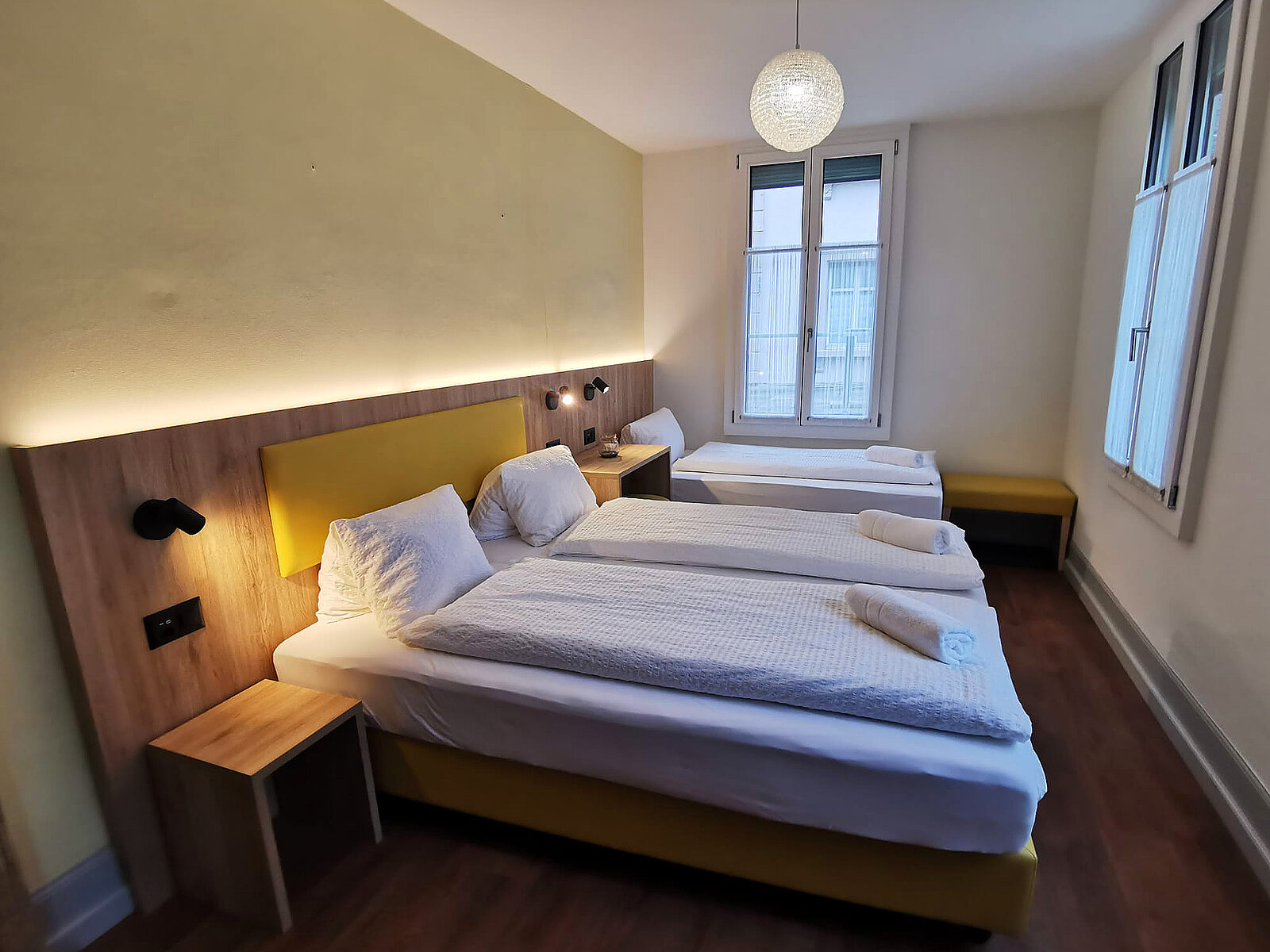 Ein helles, modernes Zimmer mit drei Einzelbetten. Die Betten sind mit weißen Bettdecken und Kopfkissen ausgestattet. An der Wand befindet sich eine stilvolle Holzverkleidung mit Beleuchtung. Rechts ist eine Bank und ein Fenster mit Blick ins Freie.
