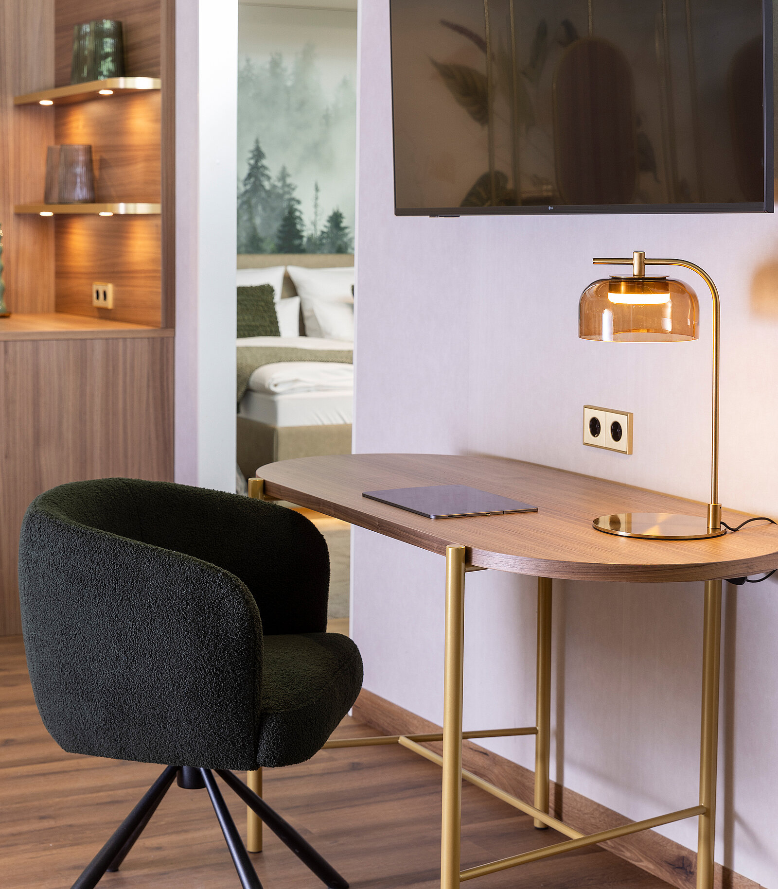A modern workspace features a stylish wooden desk with a sleek lamp and a notepad. A comfortable, dark green chair is positioned in front of the desk. In the background, a glimpse of a cozy bedroom with a soft color palette is visible.