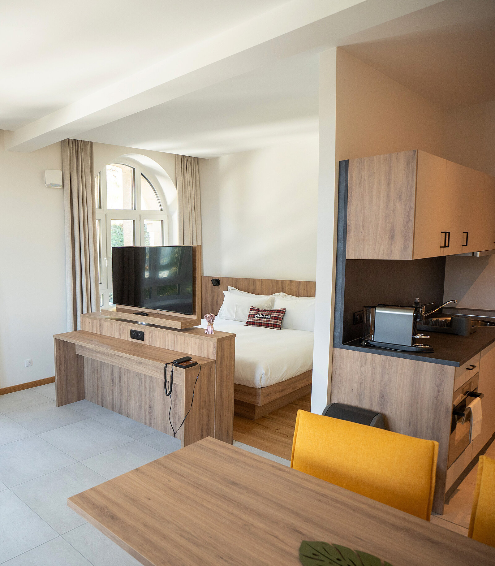 Helles Studio-Apartment mit Doppelbett, Flachbildfernseher auf Regal, kleiner moderner Küche und Esstisch mit gelben Stühlen. Große Fenster lassen natürliches Licht herein.
