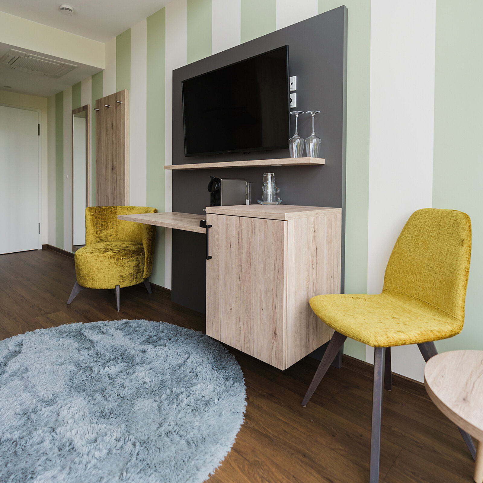 Modern eingerichtetes Hotelzimmer mit pastellgrünen Wänden, einem großen Flachbildfernseher und einem Holztisch. Zwei gelbe Stühle und ein gelber Sessel stehen um einen runden, blauen Teppich. Zwei Gläser stehen auf einem Regal neben einer Kaffeemaschine.