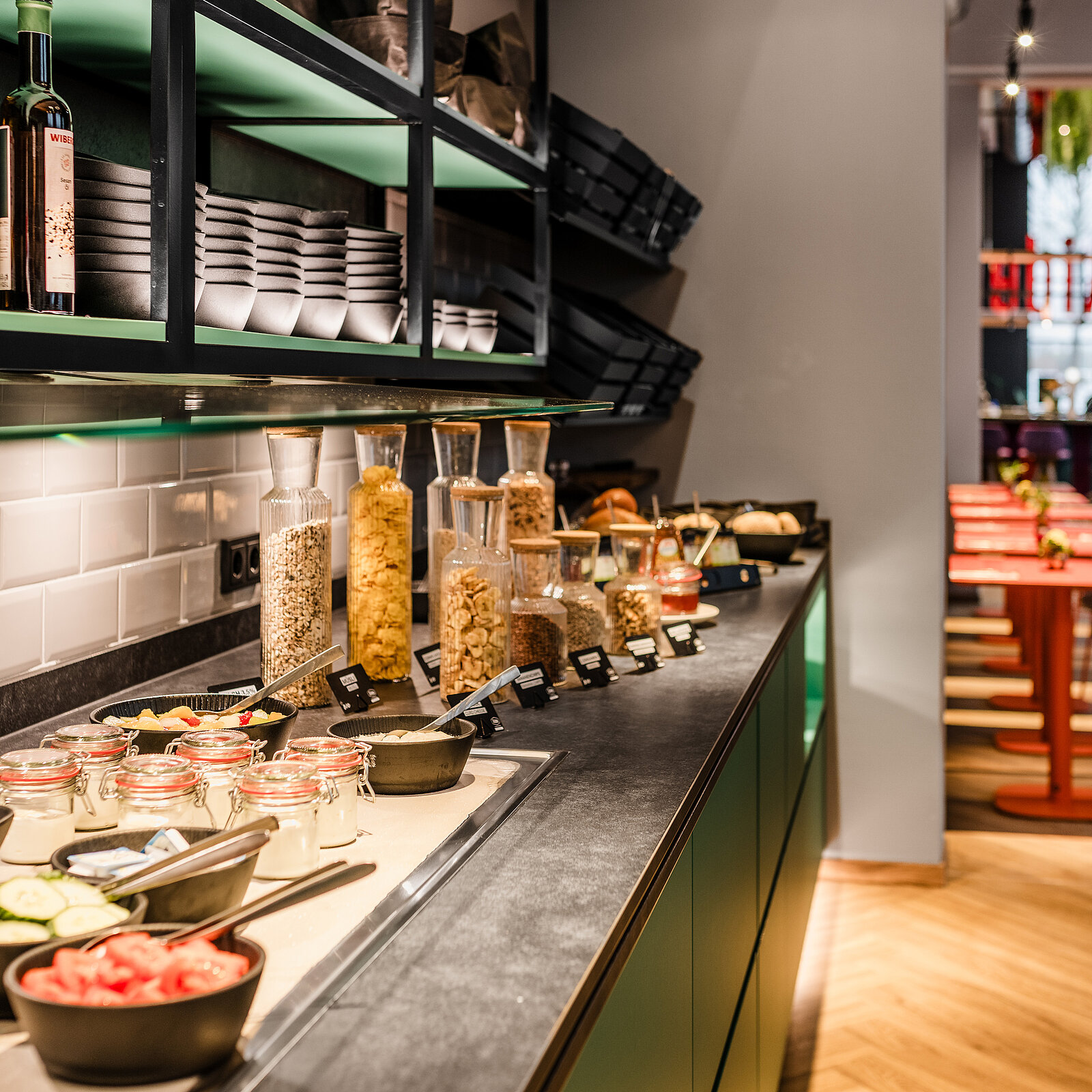 Ein Frühstücksbuffet mit Gläsern voller verschiedener Müslisorten, Flaschen, Schalen mit frischem Obst und Gemüse sowie Gewürzen auf einer langen Theke, bereit zur Selbstbedienung.