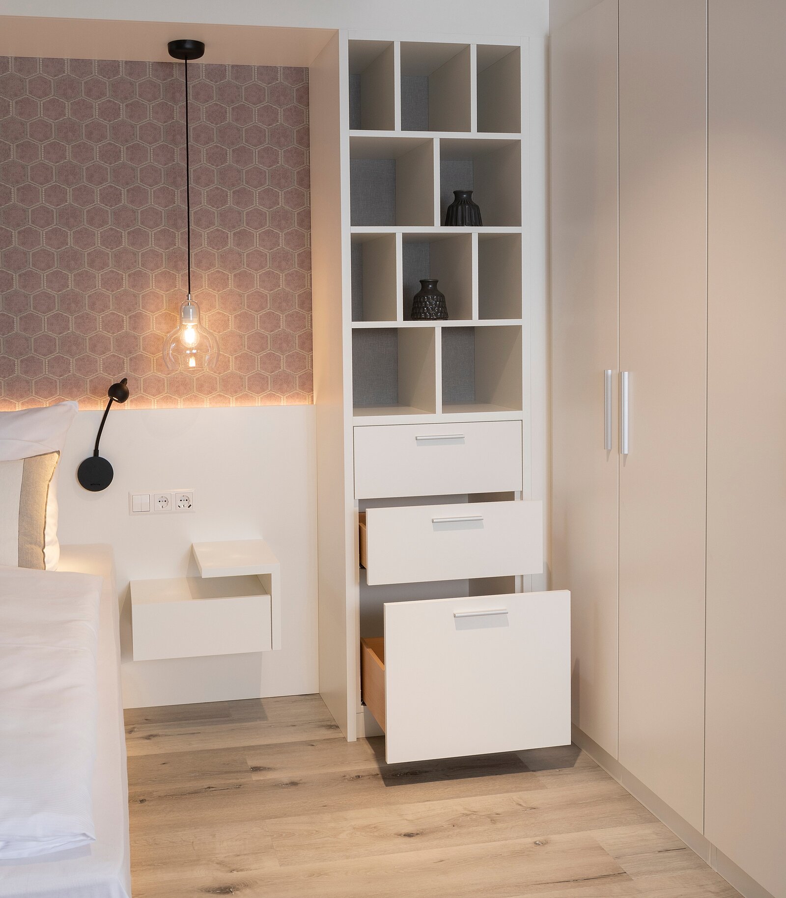 Ein moderner Schlafbereich mit einem weißen Nachttisch und einem Regal mit offenen Fächern. Neben dem Regal sind Schubladen in einem niedrigen Schrank. Die Wände sind in sanften Farben gestaltet, und eine Pendelleuchte sorgt für Beleuchtung.