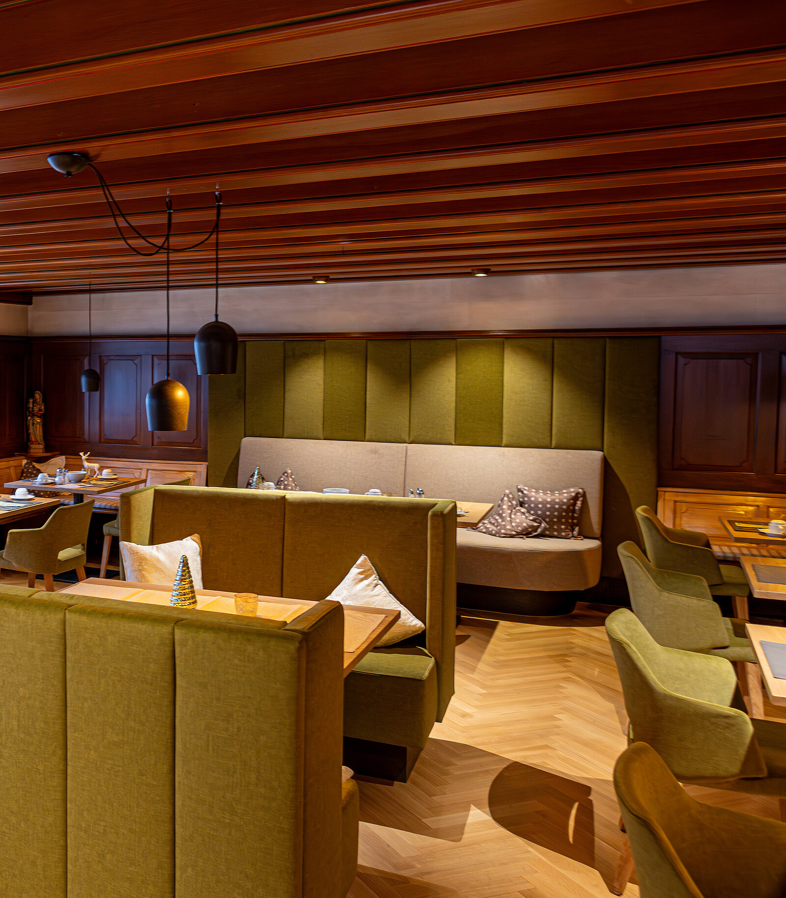 Gemütliches Restaurantinterieur mit grünen Polstersitzen und Holztischen. Die warme Beleuchtung und Holzelemente an der Decke schaffen eine einladende Atmosphäre. Dekorative Kissen ergänzen die Sitzgelegenheiten.