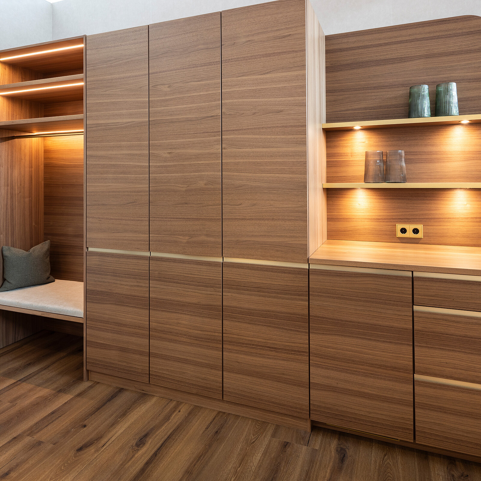 A modern room features wooden cabinetry with integrated lighting. On the left, an open shelf space is illuminated, showcasing a cozy seating area. The right side includes a closed storage unit with sleek drawers and a countertop displaying decorative items, creating a warm, inviting atmosphere.
