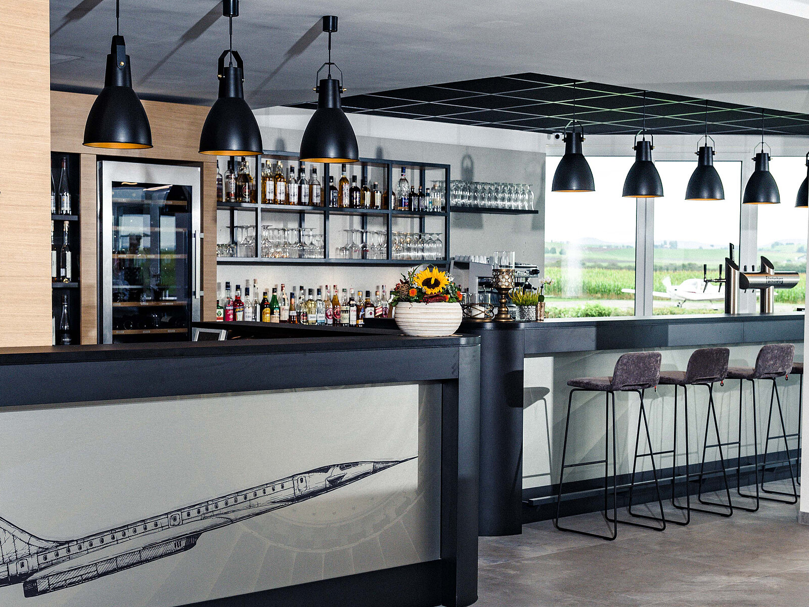 Moderne Bar mit einer Vielzahl von Getränken hinter der Theke. Über dem Tresen hängen schlichte schwarze Lampen. An der Bar stehen einige Hocker. Im Hintergrund Fenster mit Blick auf die Umgebung. Der Tresen ist mit einem grafischen Flugzeugmotiv verziert.