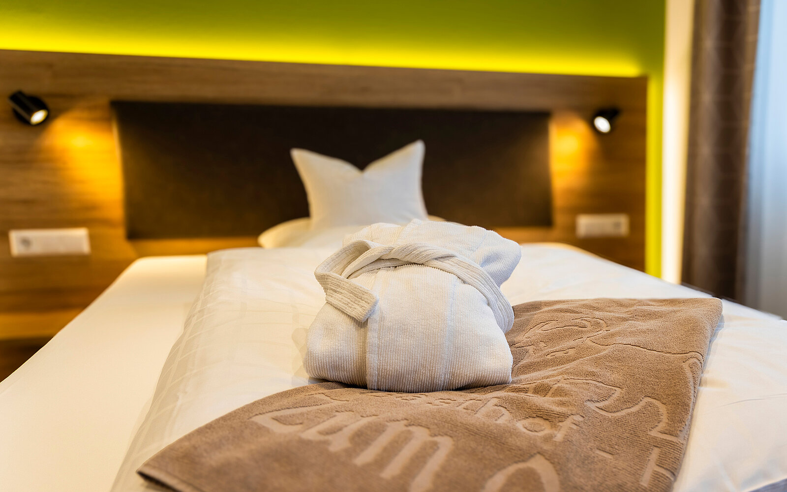 Ein gemütlich aussehendes Hotelbett mit weißen Decken und einem Dekokissen in Sternform. Auf dem Bett liegen ein zusammengelegter weißer Bademantel und ein braunes Handtuch. Die Holzwand im Hintergrund verleiht dem Raum eine warme Atmosphäre.