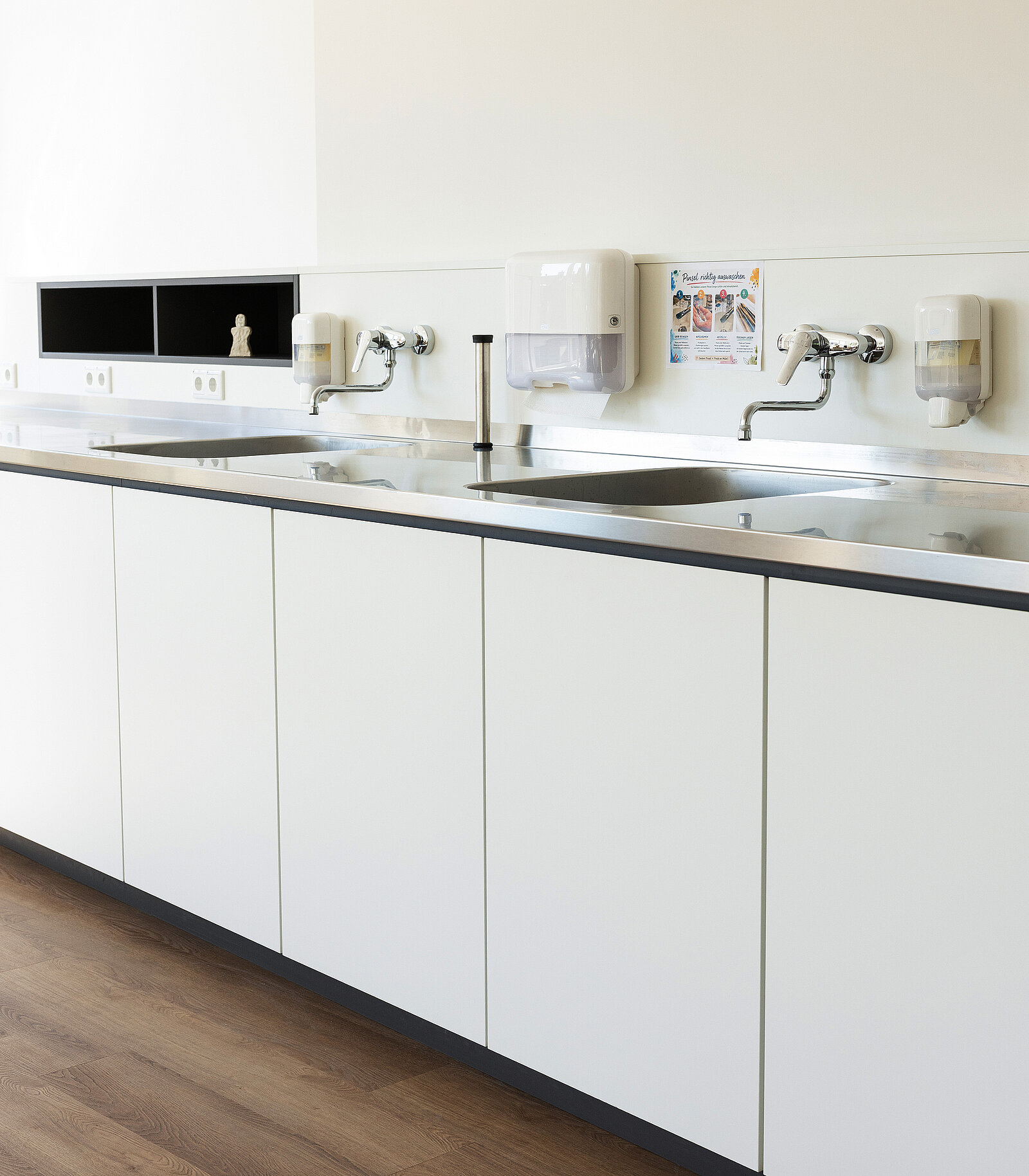 Große, weiße Küchenspüle mit zwei Edelstahlbecken, zwei Wandarmaturen und Spendern für Seife und Desinfektionsmittel. Ein Handtuchspender hängt über der Spüle. Das Design ist schlicht und modern.