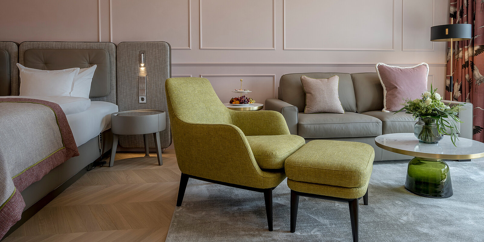 Das Bild zeigt ein modernes Hotelzimmer mit einem Bett, einem bequemen grauen Sofa und einem leuchtend grünen Sessel mit Hocker. Auf einem runden Tisch steht eine Blumenvase. Die Wände sind in sanften Farben gehalten, und das Licht schafft eine einladende Atmosphäre.