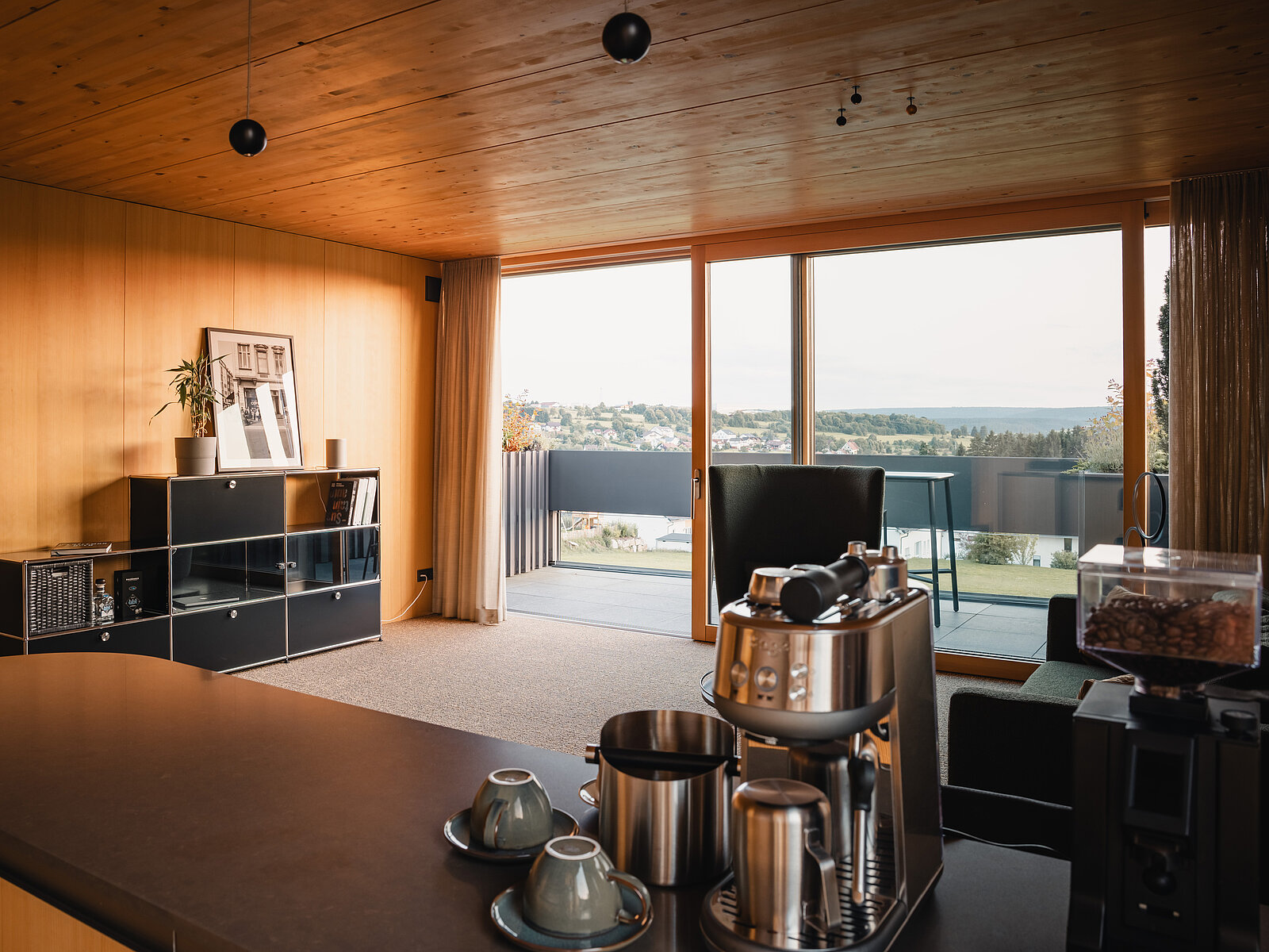 Modernes Wohnzimmer mit Holzakzenten, großem Fenster und Blick auf eine Terrasse. Im Vordergrund steht eine Espressomaschine mit Tassen. Ein Regal mit Pflanzen, Büchern und Bildern befindet sich an der Wand. Wohnlicher, offener Raum mit natürlichem Licht.
