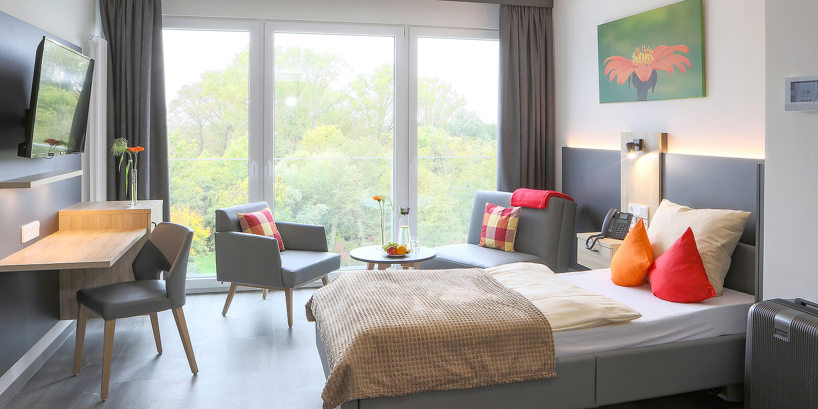 Ein modernes Hotelzimmer mit einem Einzelbett, bunten Kissen und einem Nachttisch. Vor dem Bett befindet sich ein kleiner Tisch mit einer Blumenvase. Eine Sitzecke mit zwei Stühlen und einem Tisch ist ebenfalls zu sehen. Große Fenster bieten einen Blick ins Freie.