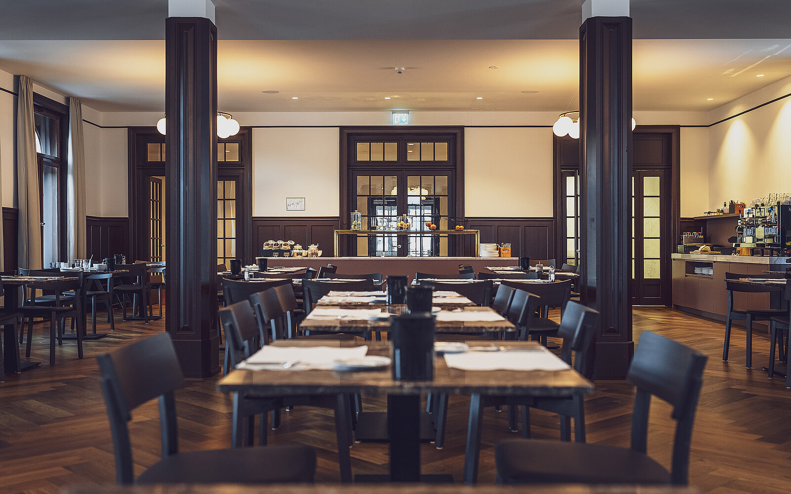 Elegantes Restaurant mit gedeckten Tischen und dunklen Holzstühlen in stilvoller, warmer Atmosphäre. Im Hintergrund befindet sich eine beleuchtete Bar. Räume sind durch große Fenster und symmetrische Anordnung der Möbel geprägt, vermittelt ein Gefühl von Ordnung und Ruhe.