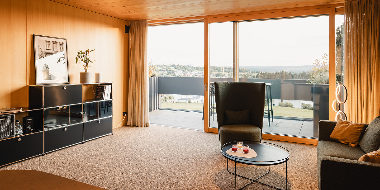Ein modernes Wohnzimmer mit Holzwänden, einem großen Fenster und Blick auf einen Balkon mit weiter Landschaft. Ein gemütlicher Sessel und ein runder Couchtisch stehen im Raum. Ein wandhohes Regal enthält Bücher und Dekorationen.