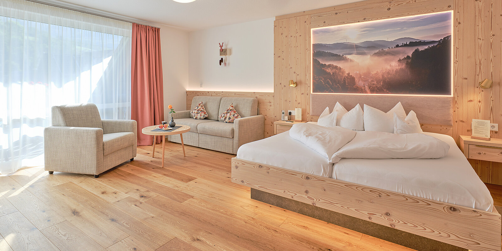 Helles Hotelzimmer mit Holzboden, einem großen Bett und einem Sofa. An der Wand über dem Bett hängt ein Bild einer Berglandschaft. Ein Sessel und ein runder Tisch mit Blumen stehen neben einem Fenster mit einem roten Vorhang.