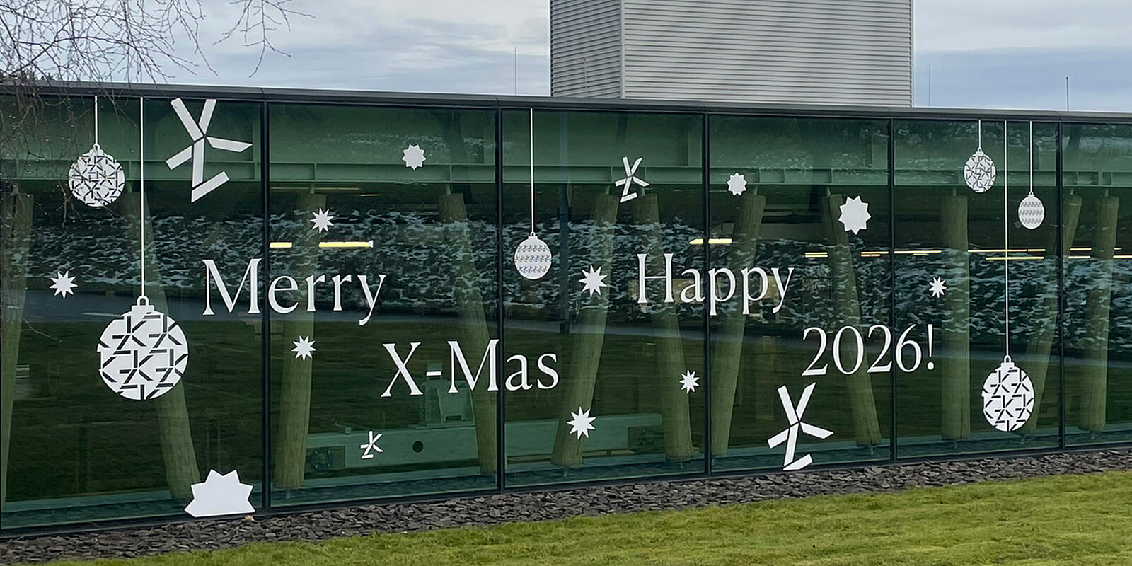 Auf einer Glaswand stehen die Worte "Merry X-Mas" und "Happy 2026!" in großen, dekorativen Buchstaben. Es sind auch weiße Weihnachtsbaumkugeln und Schneeflocken als dekorative Elemente zu sehen, die festliche Stimmung vermitteln.