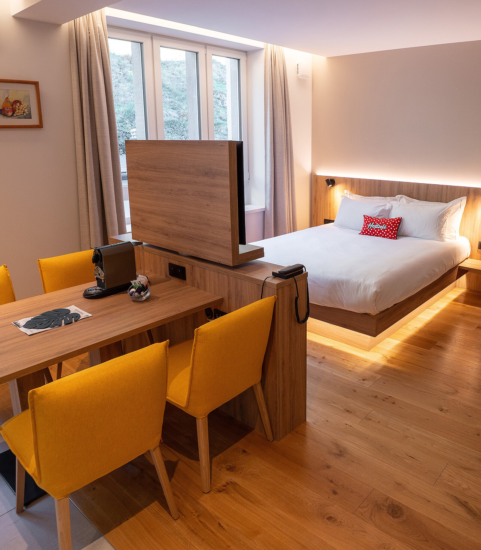 Gemütliches Hotelzimmer mit Holzboden, großem Bett und hellen Kissen. Ein Tisch mit vier gelben Stühlen steht gegenüber einem Holz-Schreibtisch mit einer Kaffeemaschine. Großer Fernseher auf dem Tisch vor dem Bett. Fenster lassen Tageslicht herein.