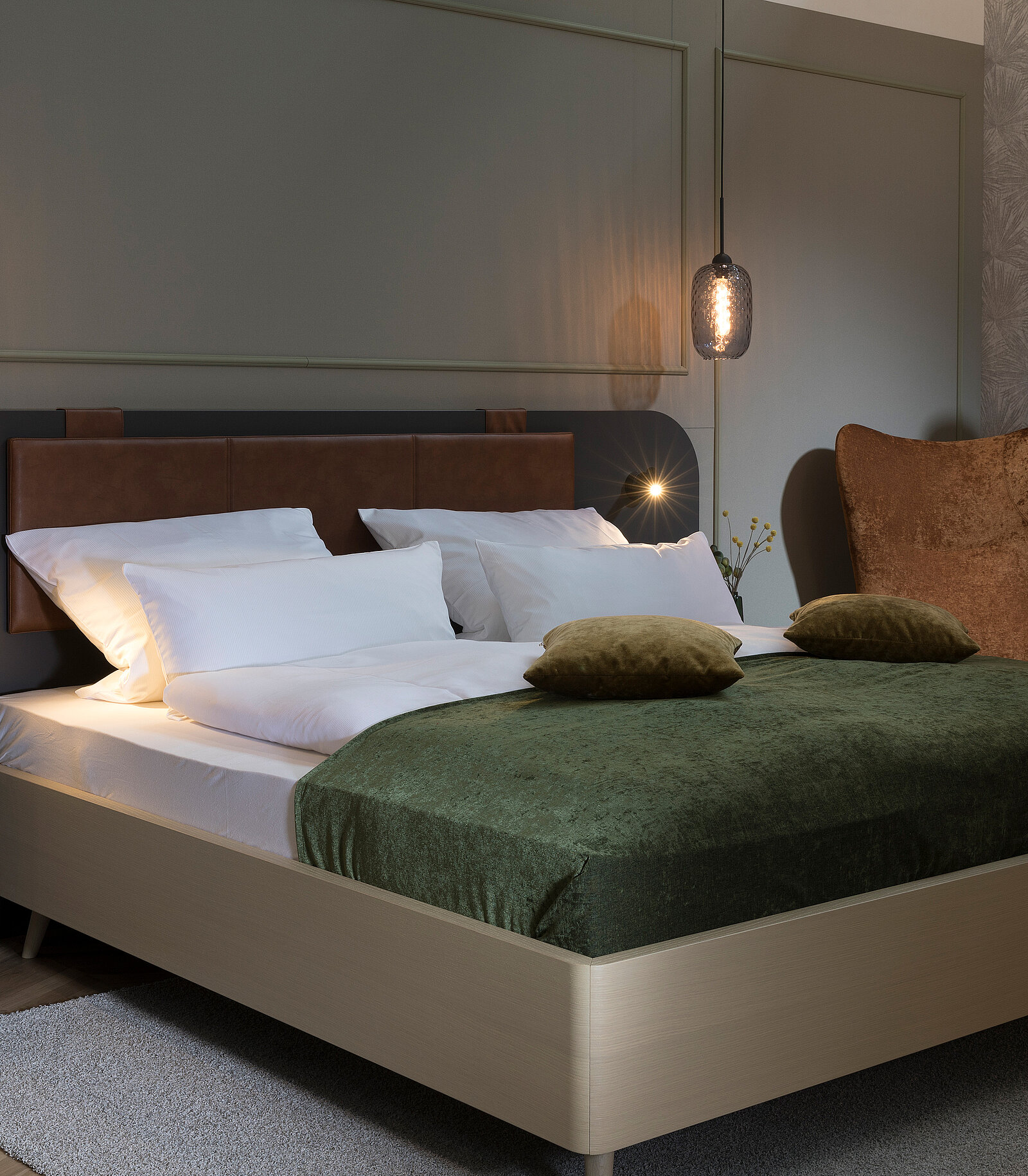 Das Bild zeigt ein modernes Schlafzimmer mit einem bequemen Bett, das mit weißen Kissen und einer grünen Decke bedeckt ist. An der Wand hängt eine stilvolle Lampe, und die Wände sind in neutralen Farbtönen gestaltet, was eine gemütliche Atmosphäre schafft.