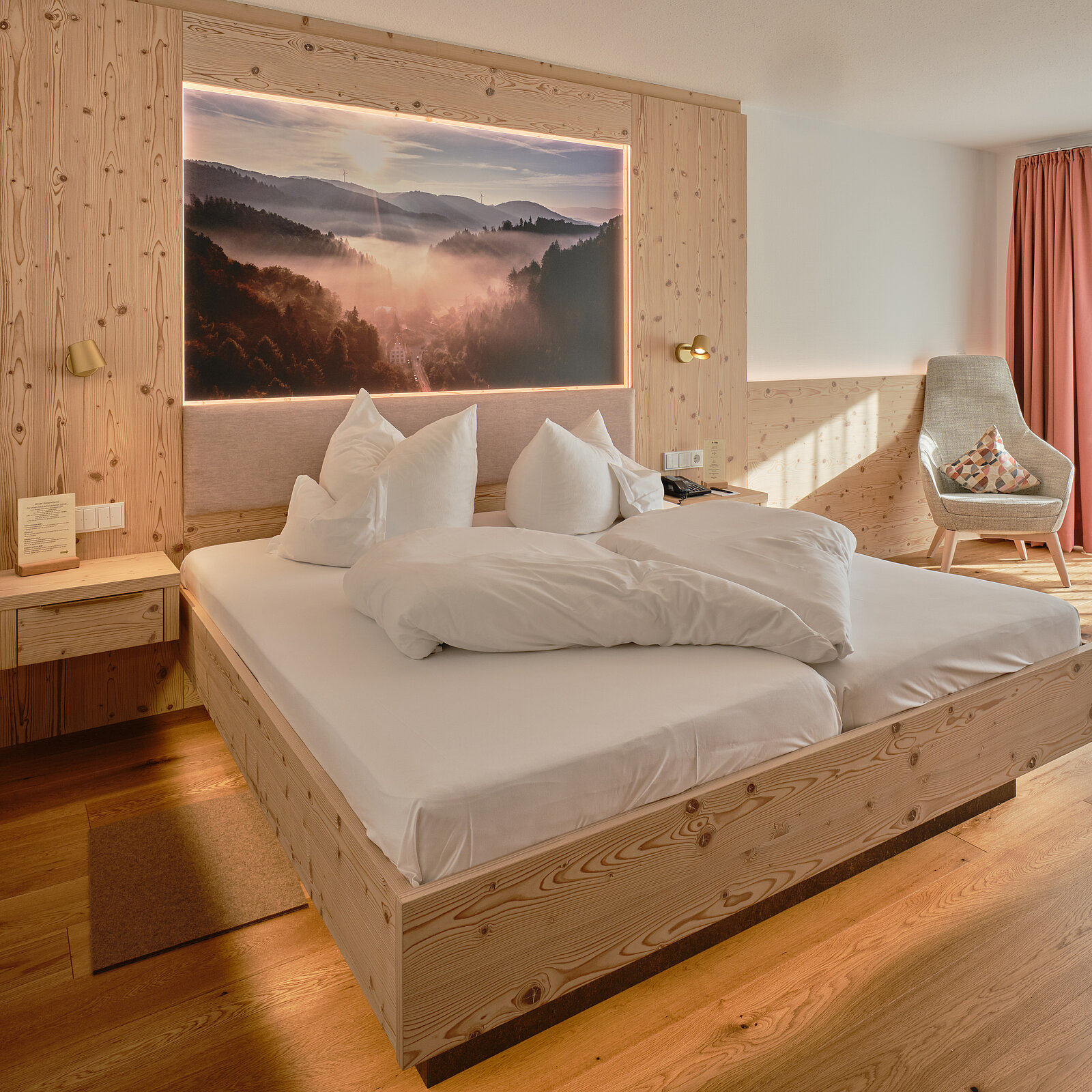 Gemütliches Hotelzimmer mit einem großen Holzbett, das mit weißen Kissen und Decken ausgestattet ist. An der Wand hängt ein beleuchtetes Bild mit Berglandschaft. Rechts vom Bett steht ein Sessel neben einem Fenster mit einem Vorhang. Holzverkleidung an Wänden und Boden.