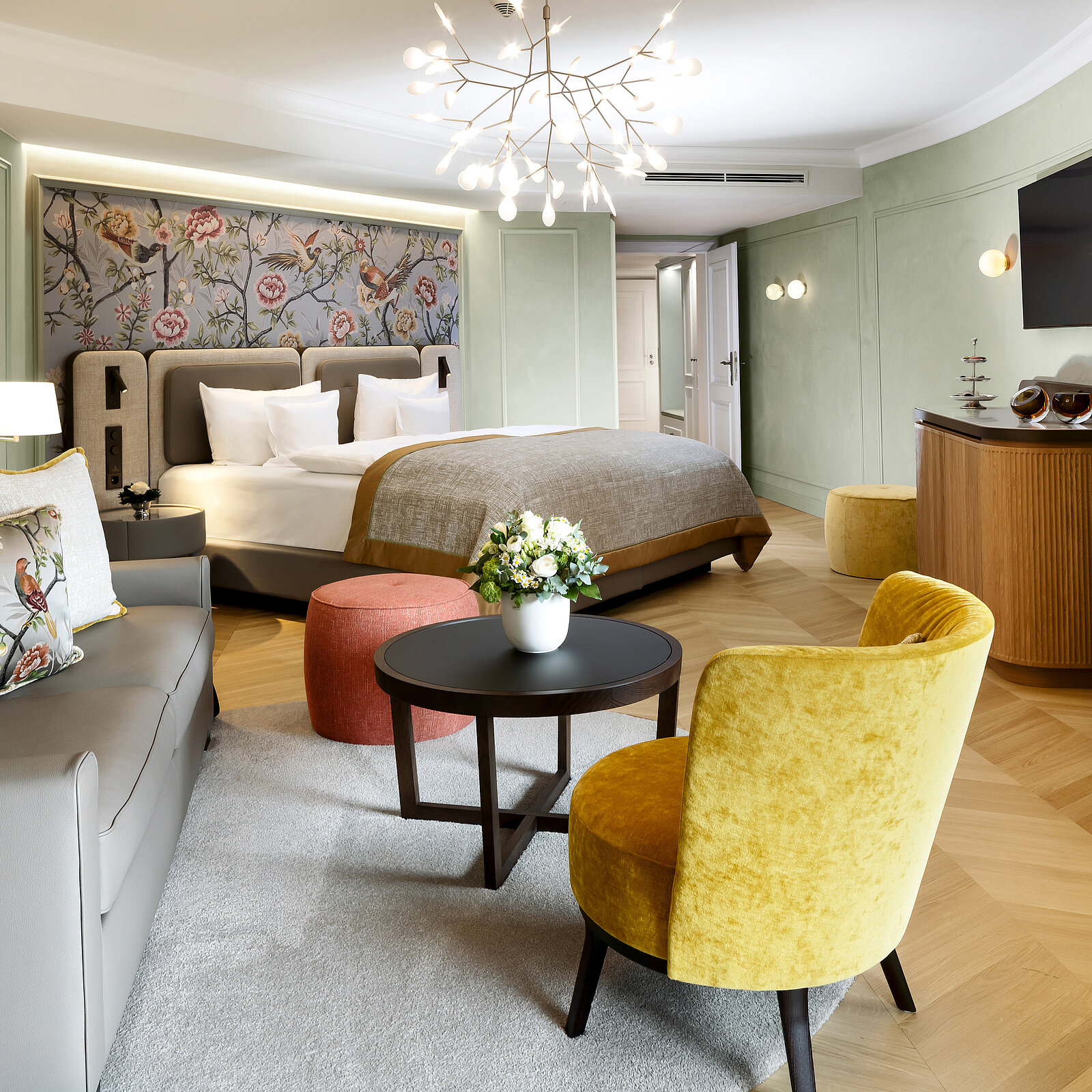 Ein elegant eingerichtetes Hotelzimmer mit einem großen Doppelbett, dekorativem Kopfteil, einem Sofa, einem runden Couchtisch mit Blumen und einem gelben Sessel. Die Wände sind hellgrün, es gibt einen modernen Kronleuchter und einen großen Fernseher an der Wand.