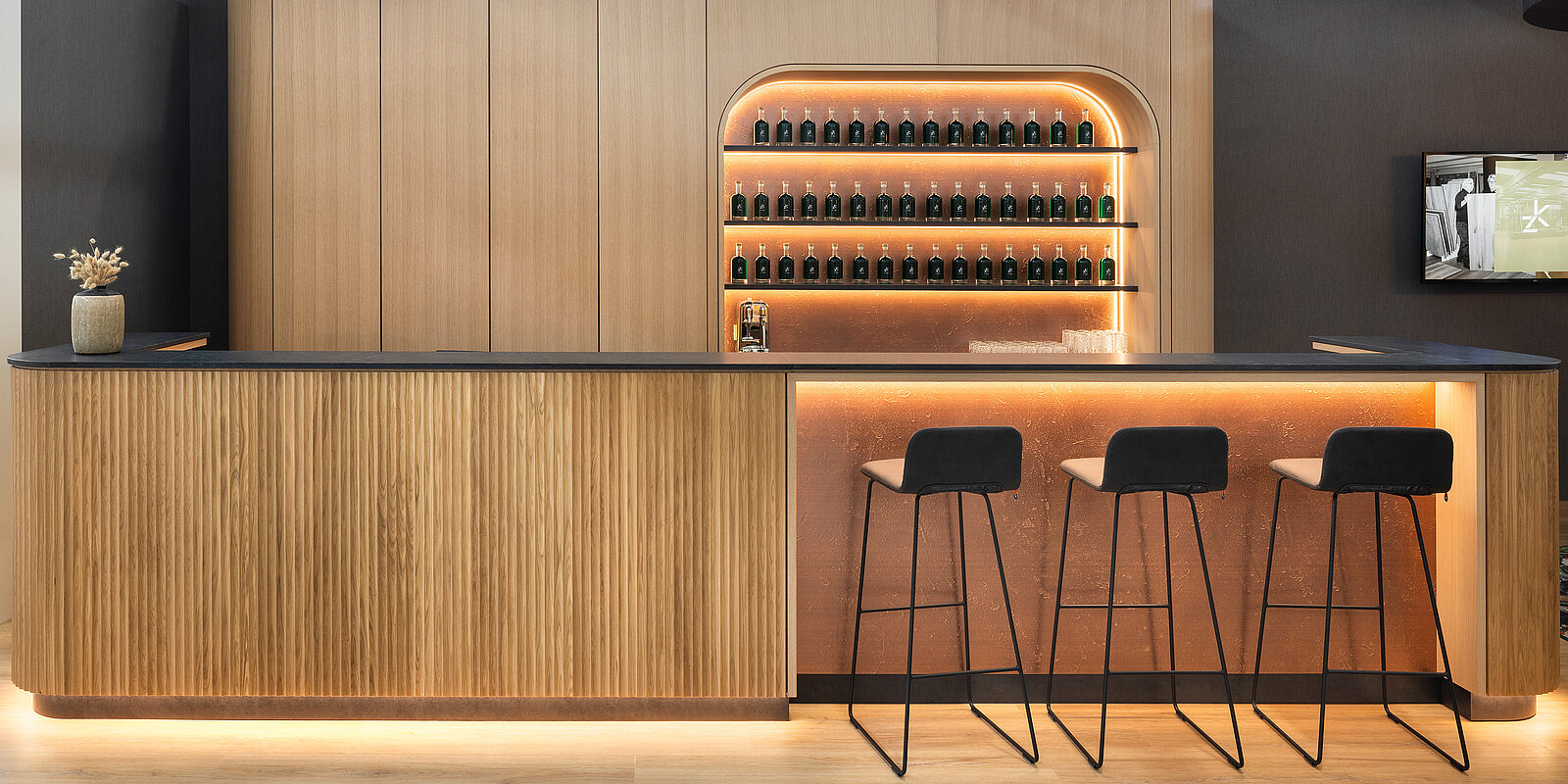 A modern bar featuring a curved wooden counter with three black stools. Behind the bar, a softly illuminated shelf displays green bottles. The design has a warm and inviting ambiance, highlighted by warm lighting and a sleek finish.