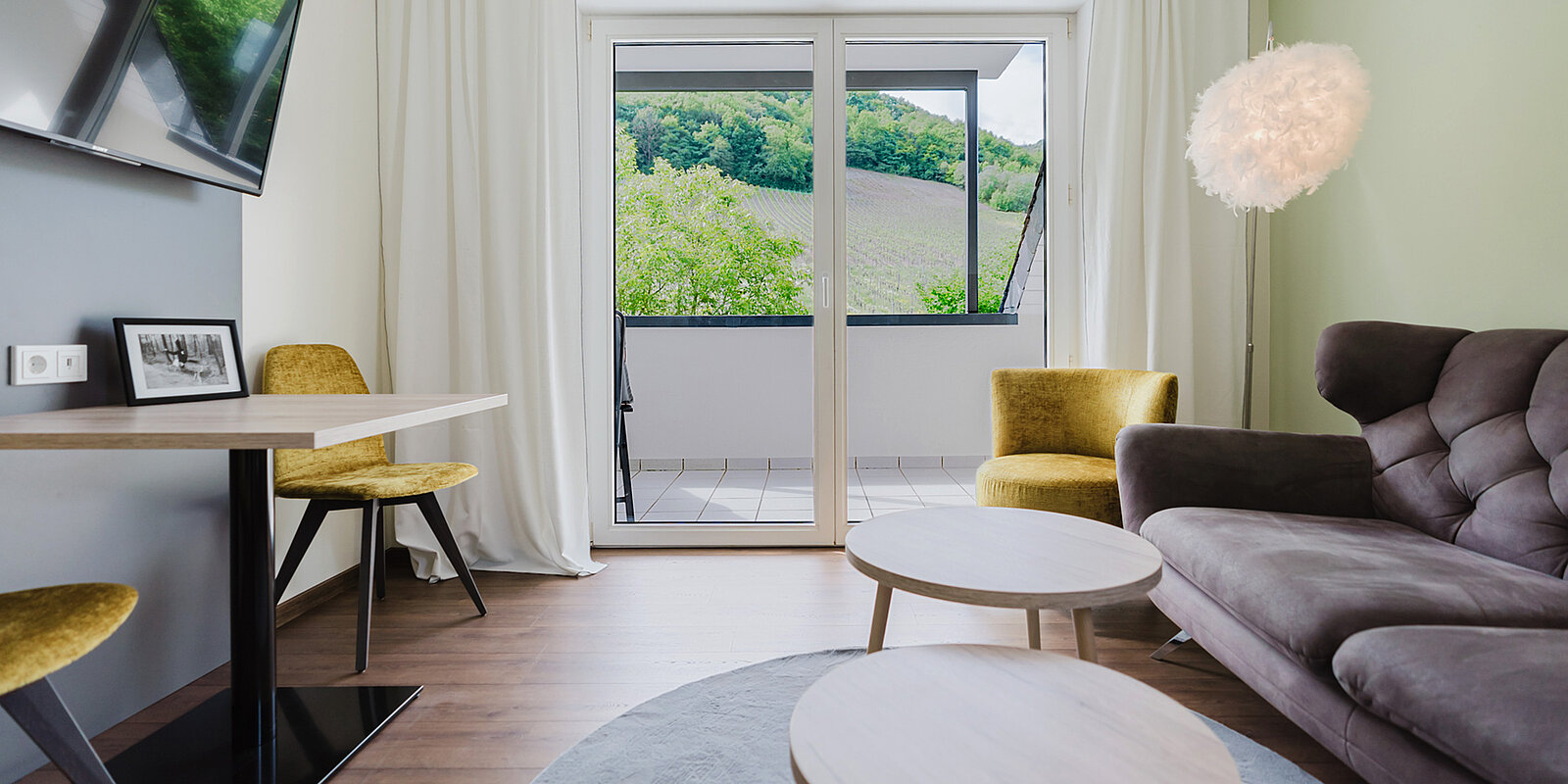 Ein modernes Wohnzimmer mit einem hellen Sofa, zwei gelben Sesseln, einem runden Holztisch und einem Tisch vor großen Fenstertüren. Die Türen führen auf einen Balkon mit Blick auf grüne Hügel. Eine dekorative Lampe hängt von der Decke.